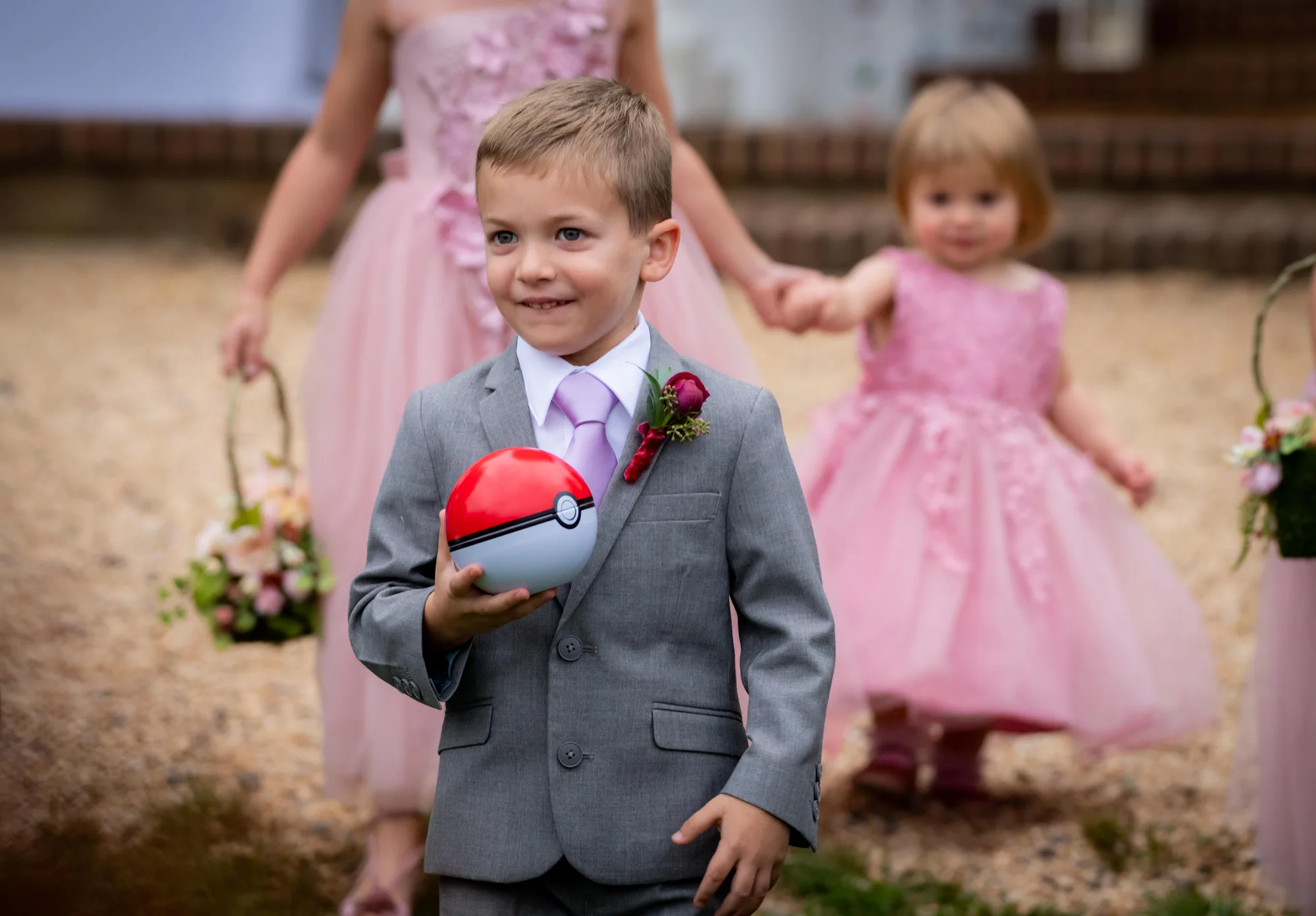 Smiling ring bearer in gray suit carries a Poké Ball down the aisle at a wedding ceremony