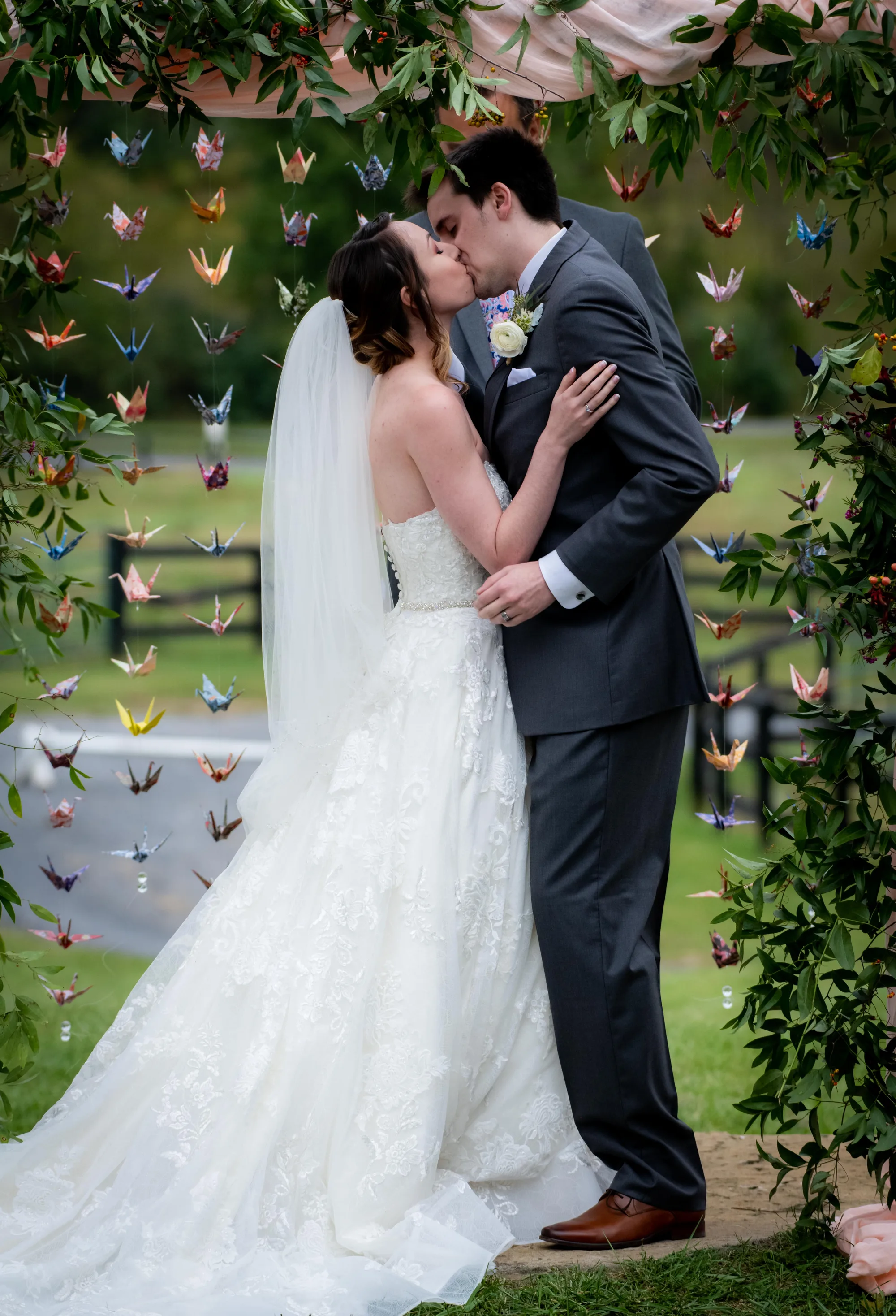 Bride and groom share first kiss under floral arch adorned with colorful origami cranes at Rixey Manor