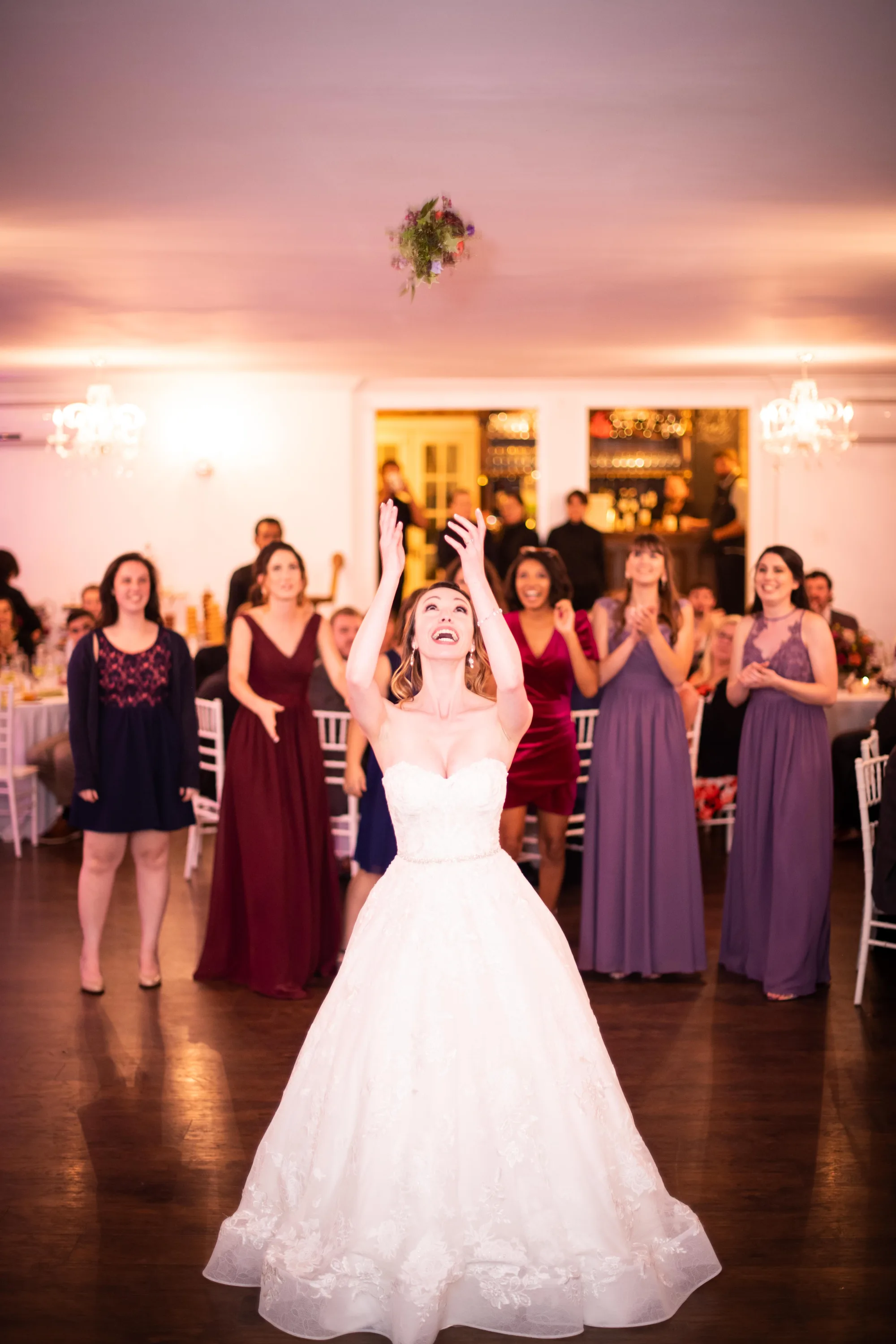 Bride tosses bouquet to guests in warmly lit reception hall as crowd watches with excitement