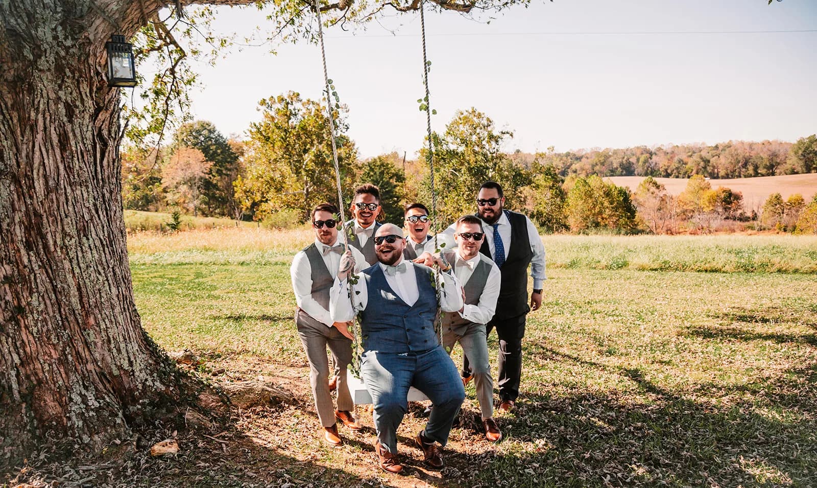 Groom sits on tree swing surrounded by laughing groomsmen on sunny Rixey Manor grounds in fall