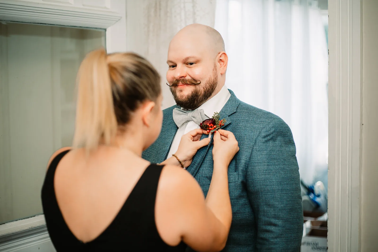 Woman in black dress adjusts groom's bow tie in front of mirror indoors