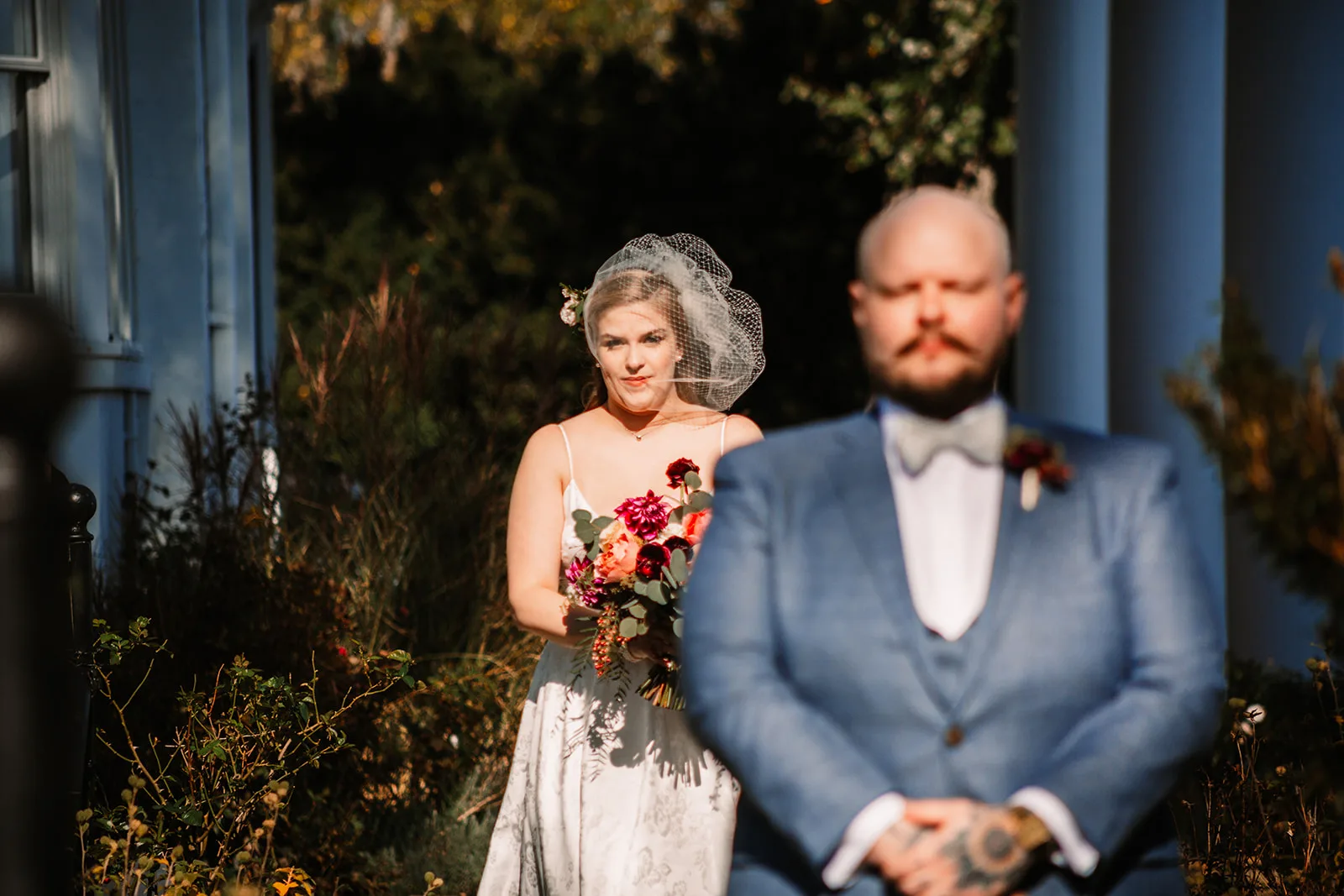 Bride in white dress with red bouquet and groom in blue suit pose outside manor columns at dusk