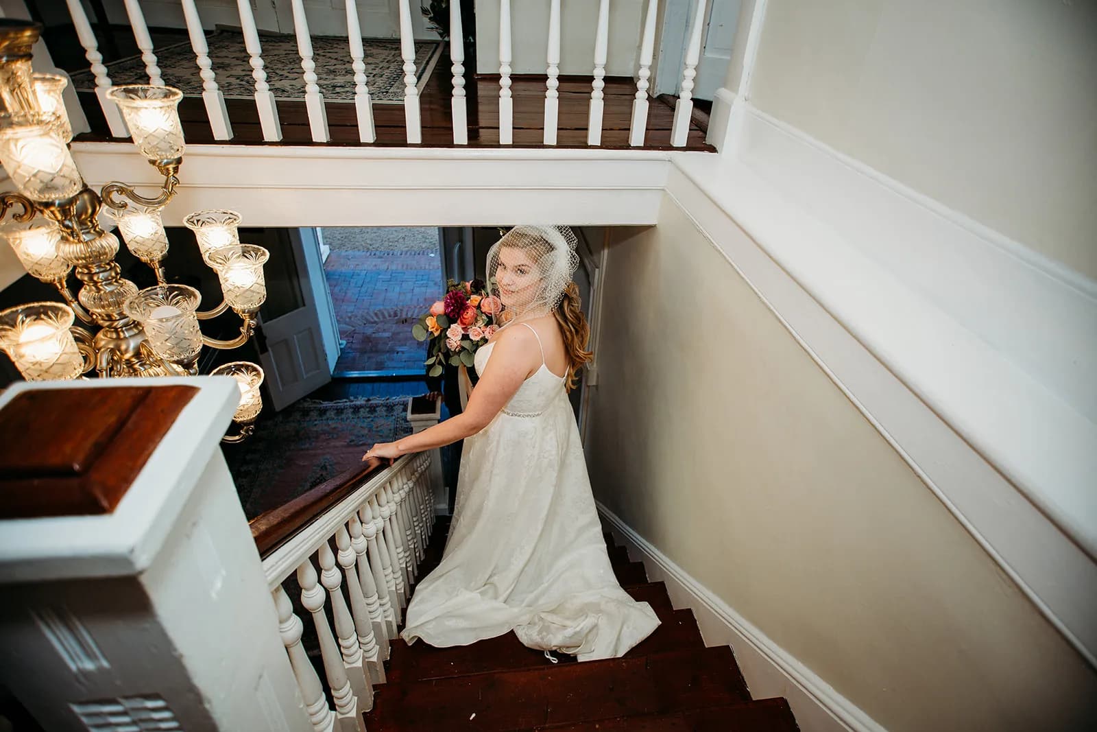 Bride descending grand staircase at Rixey Manor holding colorful bouquet, crystal chandelier overhead