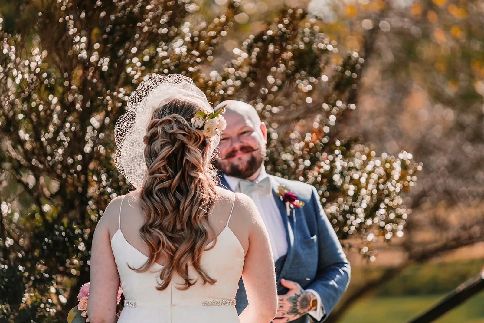 Bride and groom share a warm moment outdoors, groom smiling in blue suit with boutonniere at golden hour