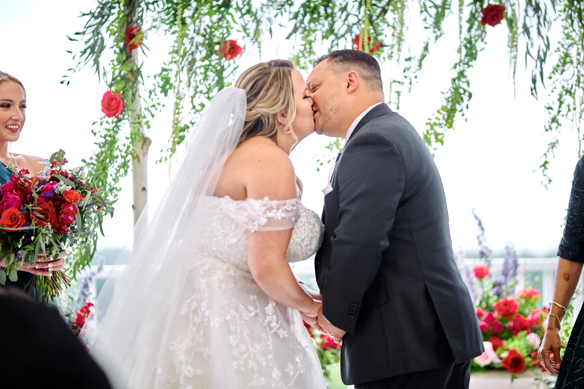 Bride and groom share first kiss under floral arch at outdoor ceremony, bridesmaid holding red bouquet nearby
