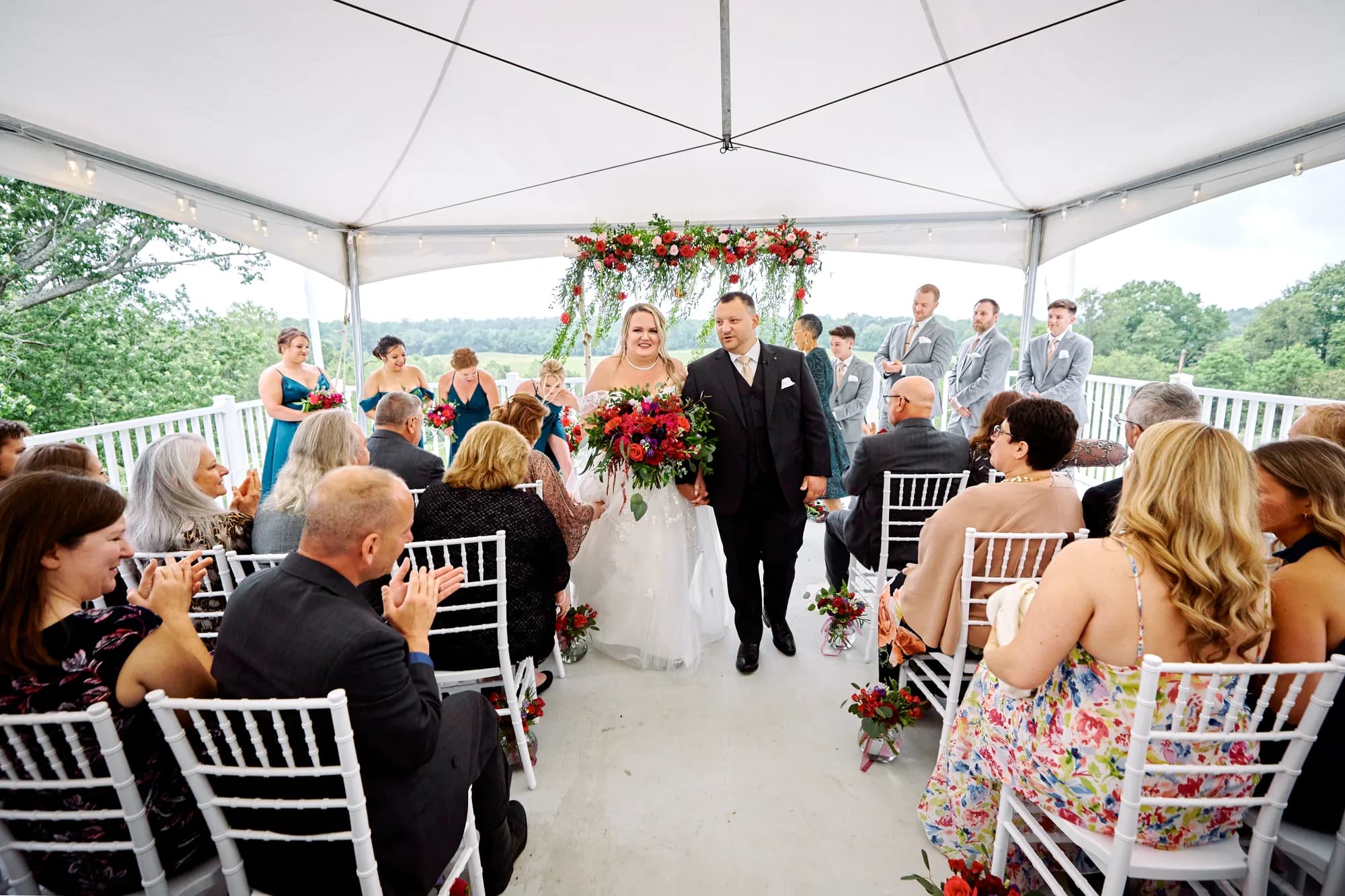Newlyweds walk the aisle under Rixey Manor's tent ceremony pavilion with red floral arch and guests applauding