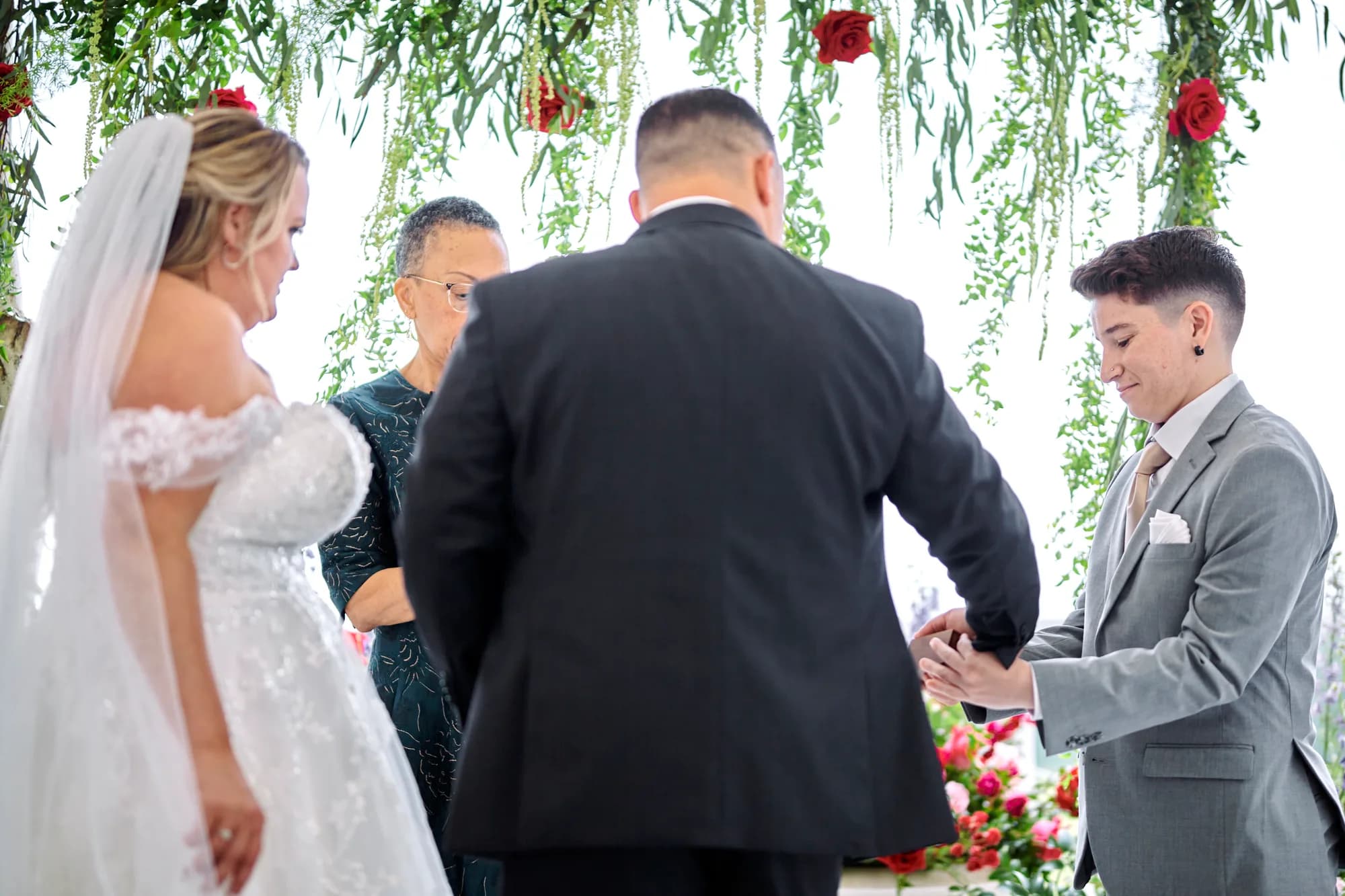 Ring exchange moment at floral arch ceremony with bride in white gown and partner in gray suit