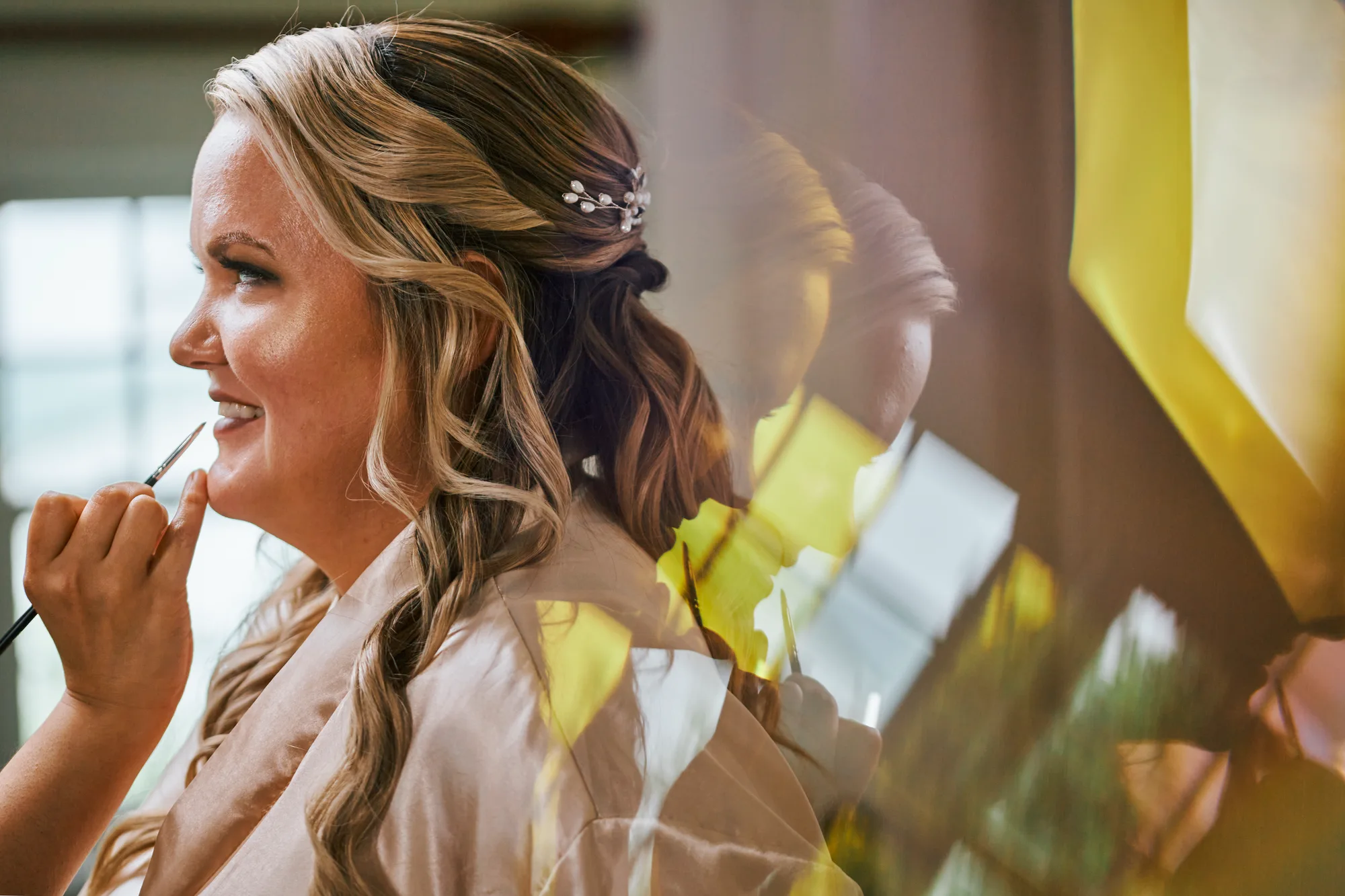 Smiling bride getting makeup applied, wearing a satin robe with floral hair piece, glowing with anticipation