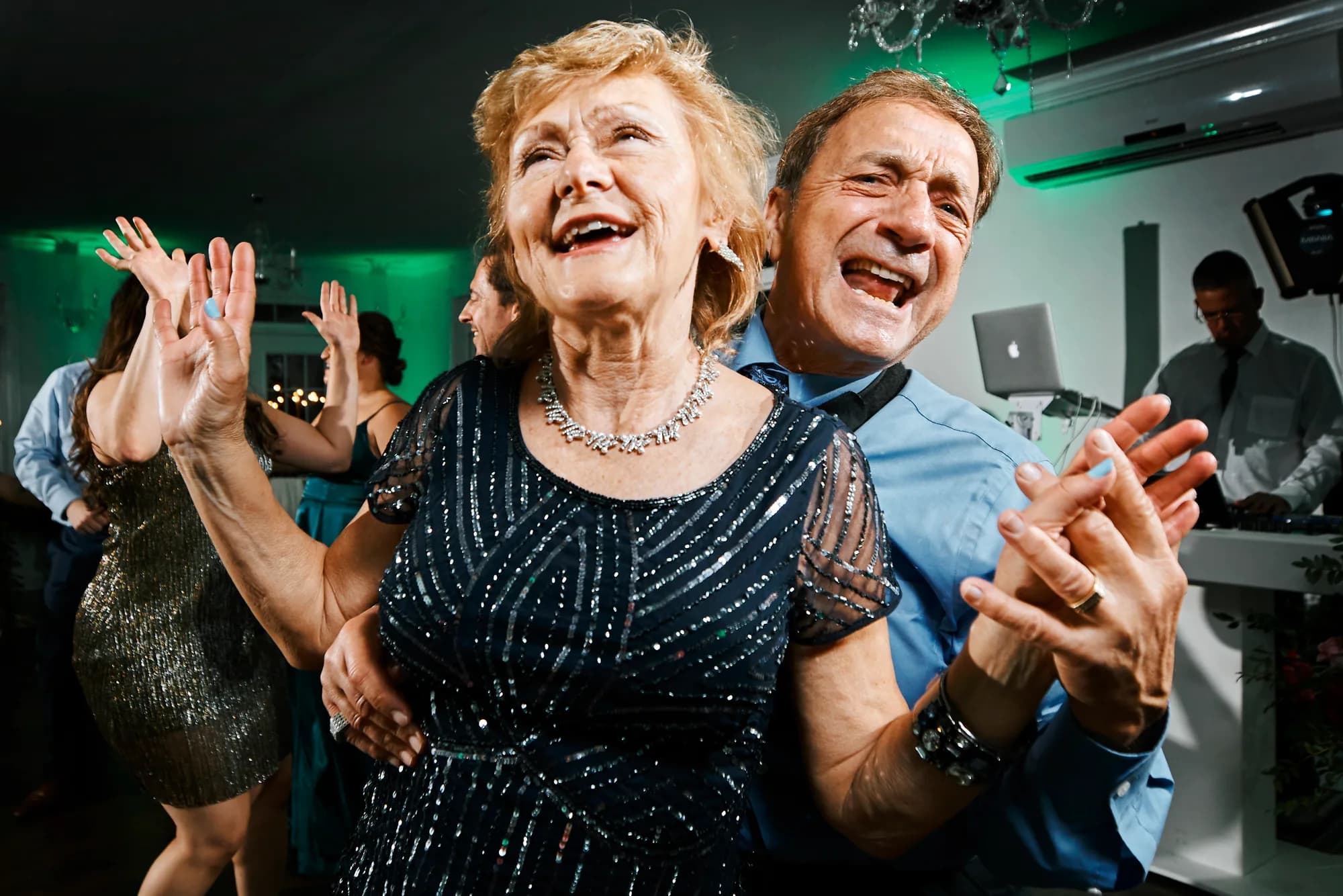 Elderly couple laughing and dancing joyfully on the reception dance floor with DJ visible in background