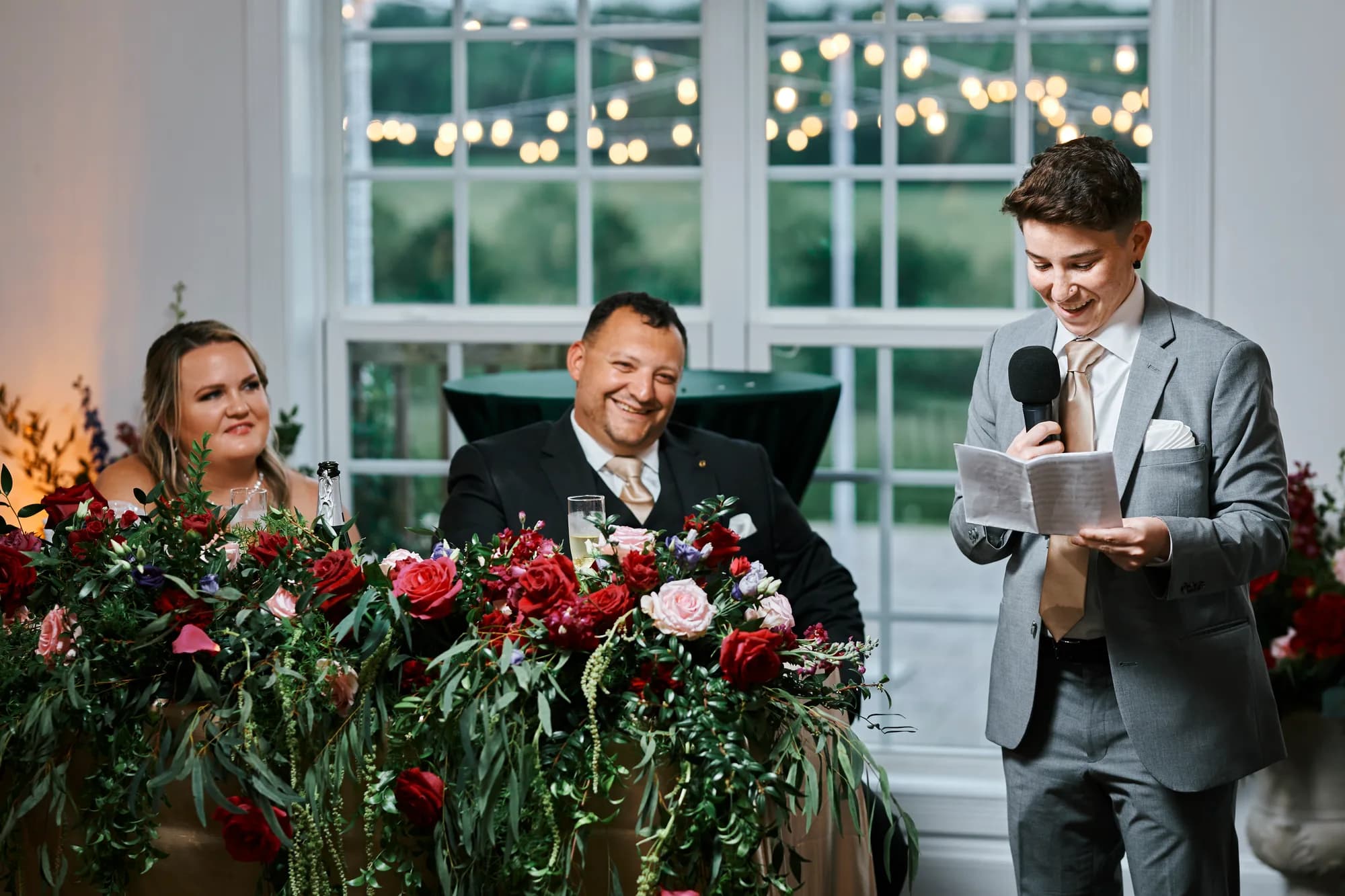 Best man reads speech at sweetheart table adorned with red and pink florals as couple smiles warmly