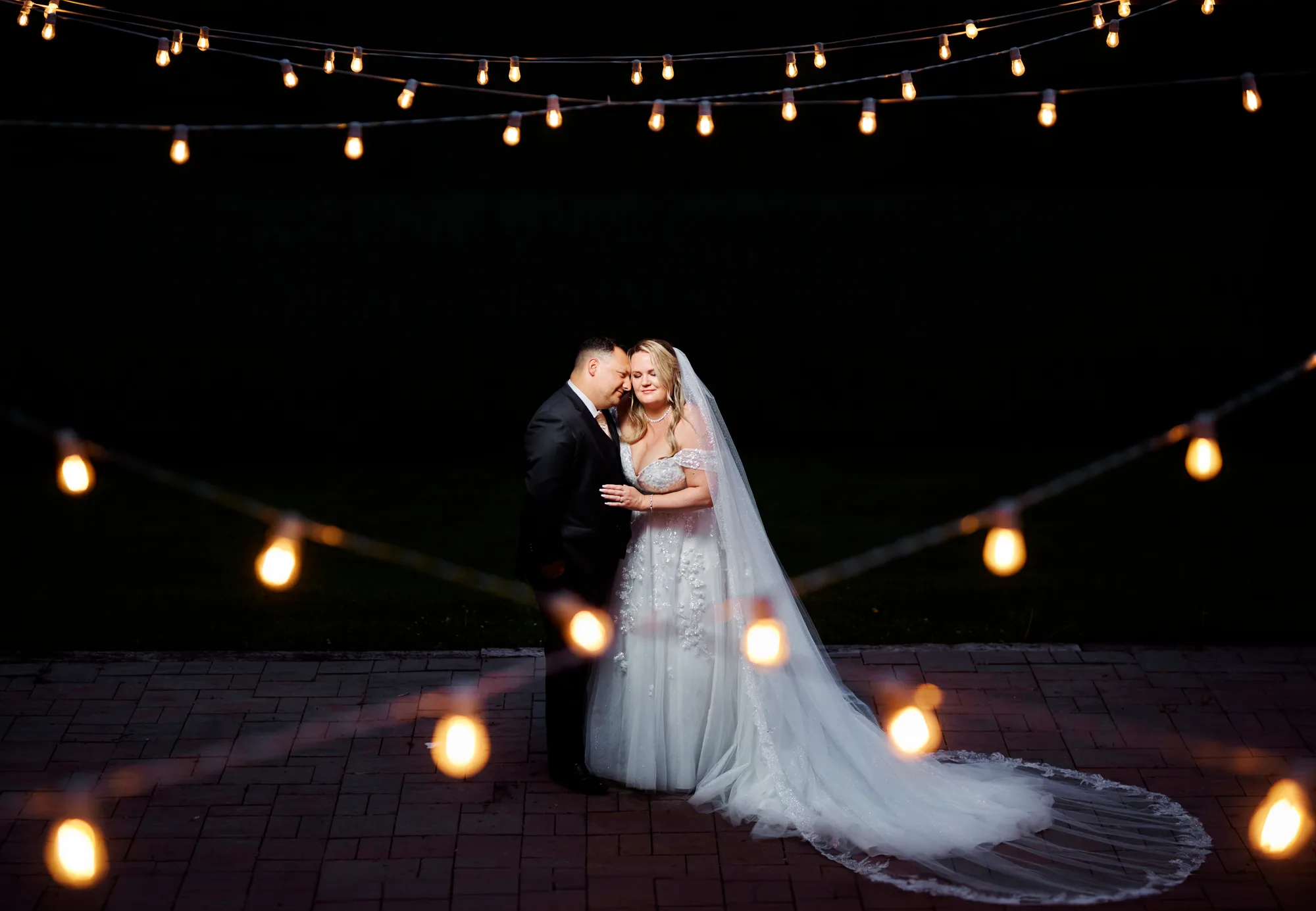 Bride and groom share a tender moment under glowing string lights on a brick terrace at night