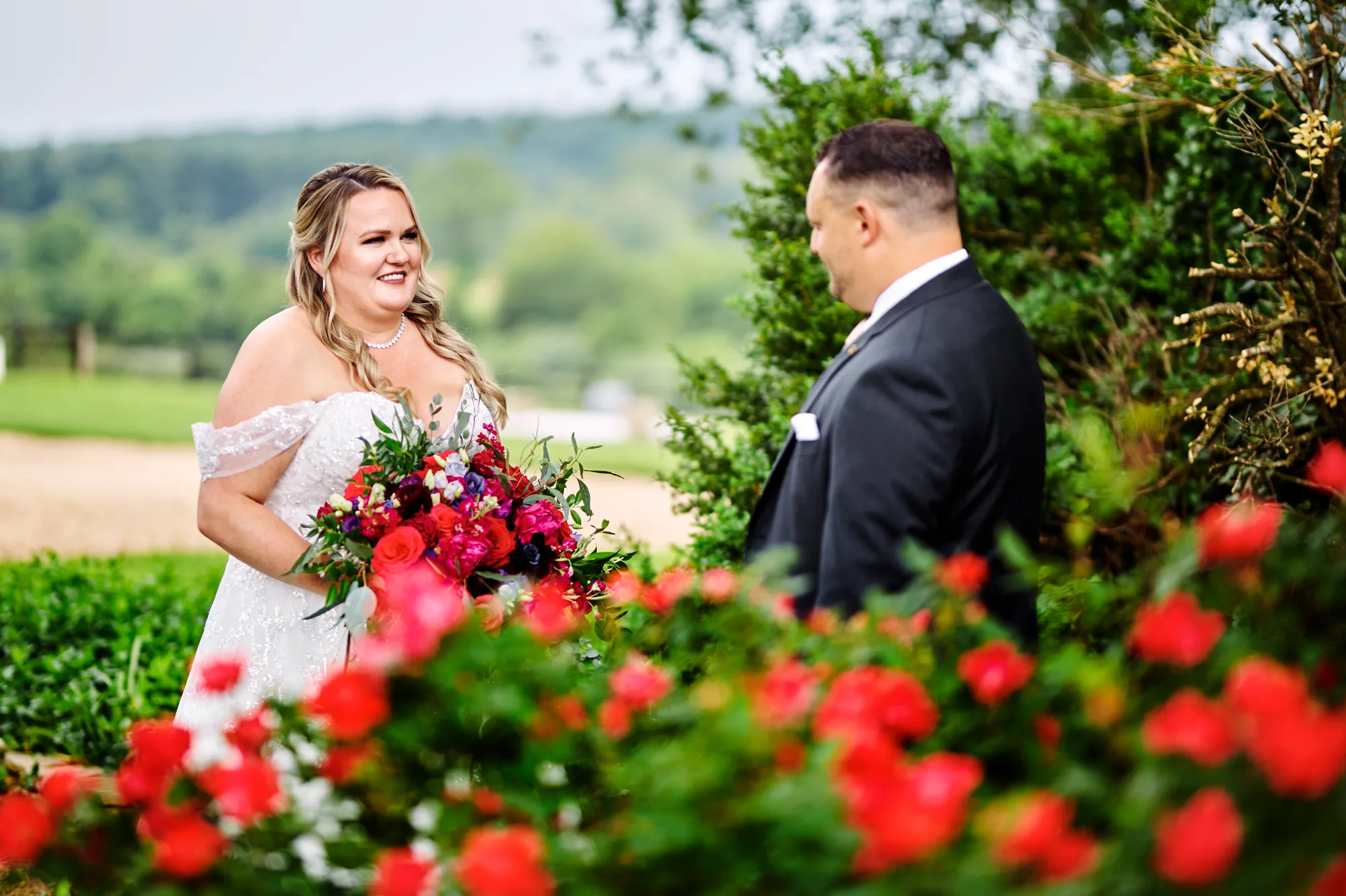 Bride smiles at groom during first look among red blooms at Rixey Manor's lush garden grounds