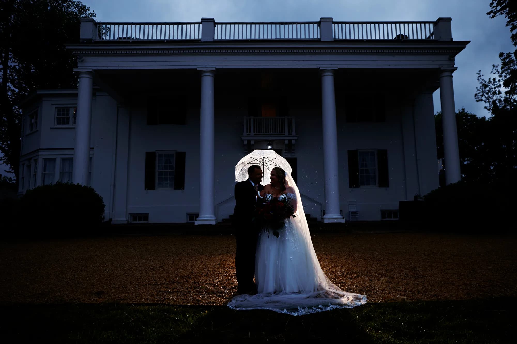 Bride and groom silhouetted under glowing umbrella at dusk before Rixey Manor's grand white-columned facade