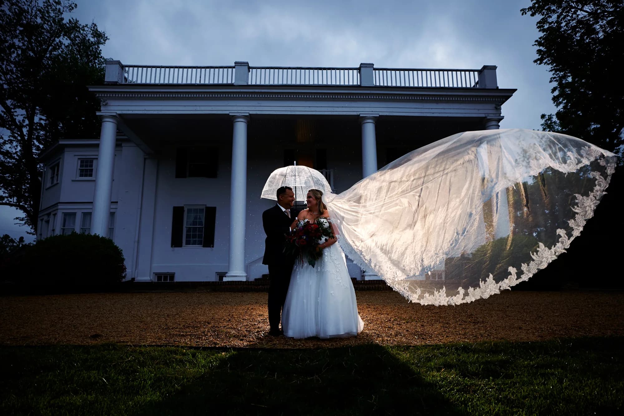 Bride and groom share an intimate moment under a clear umbrella as her veil billows before Rixey Manor's columned facade at dusk.
