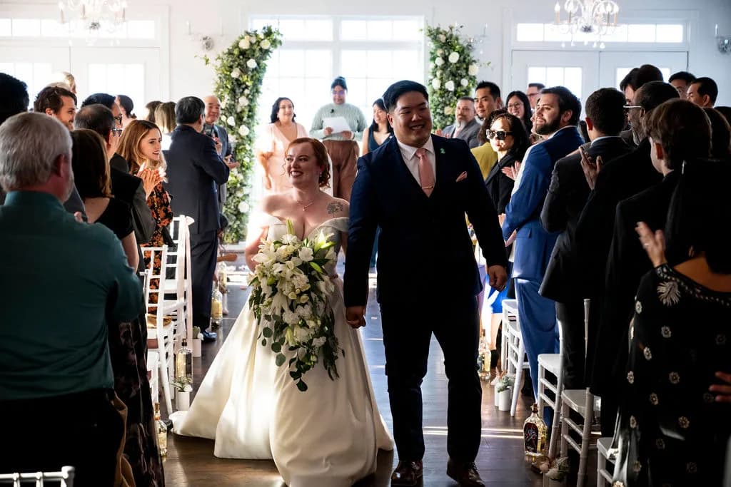 Beaming newlyweds walk the aisle at Rixey Manor ceremony room, guests applauding, floral arch behind them