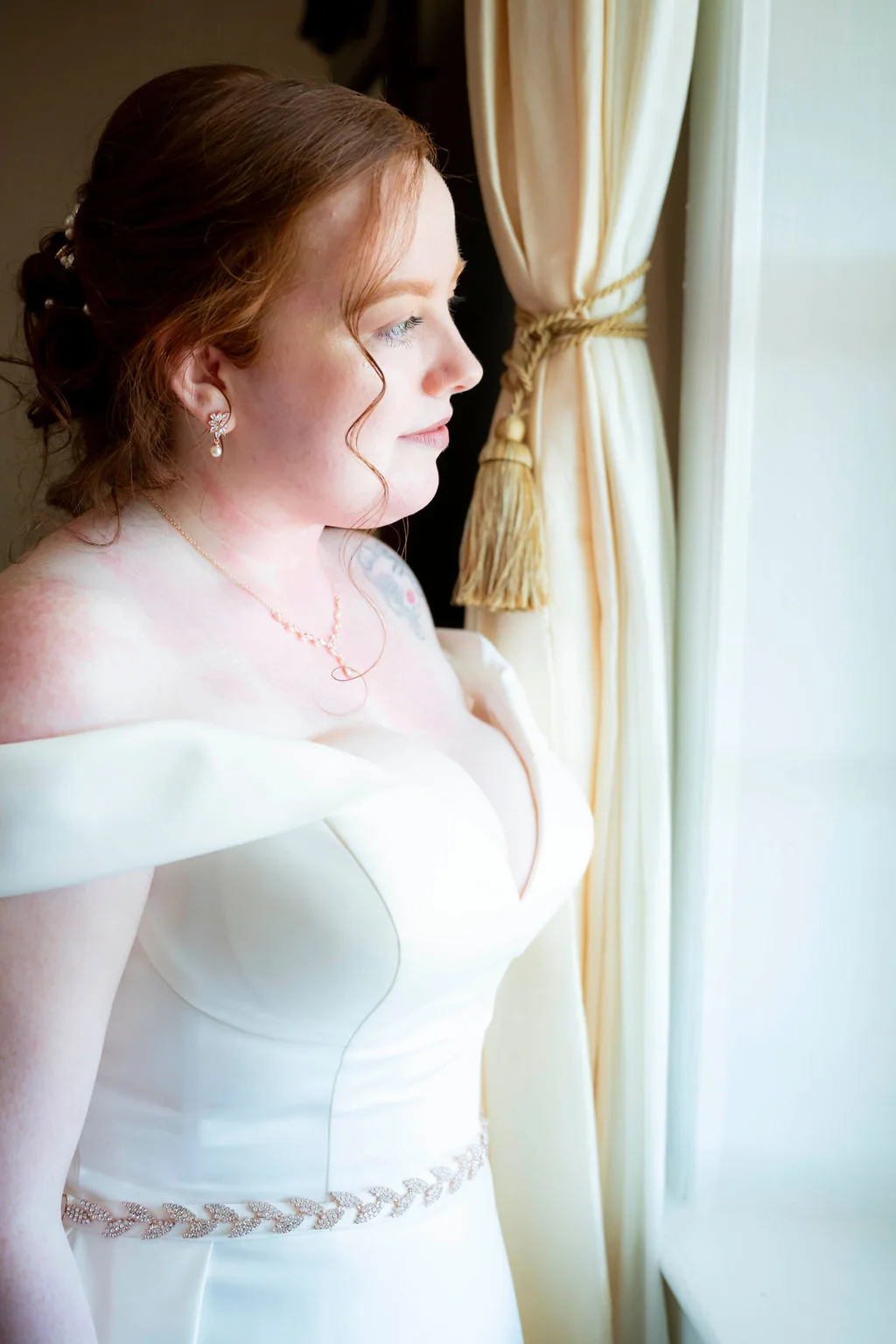 Bride in off-shoulder gown gazes pensively out a window with draped cream curtains before her wedding