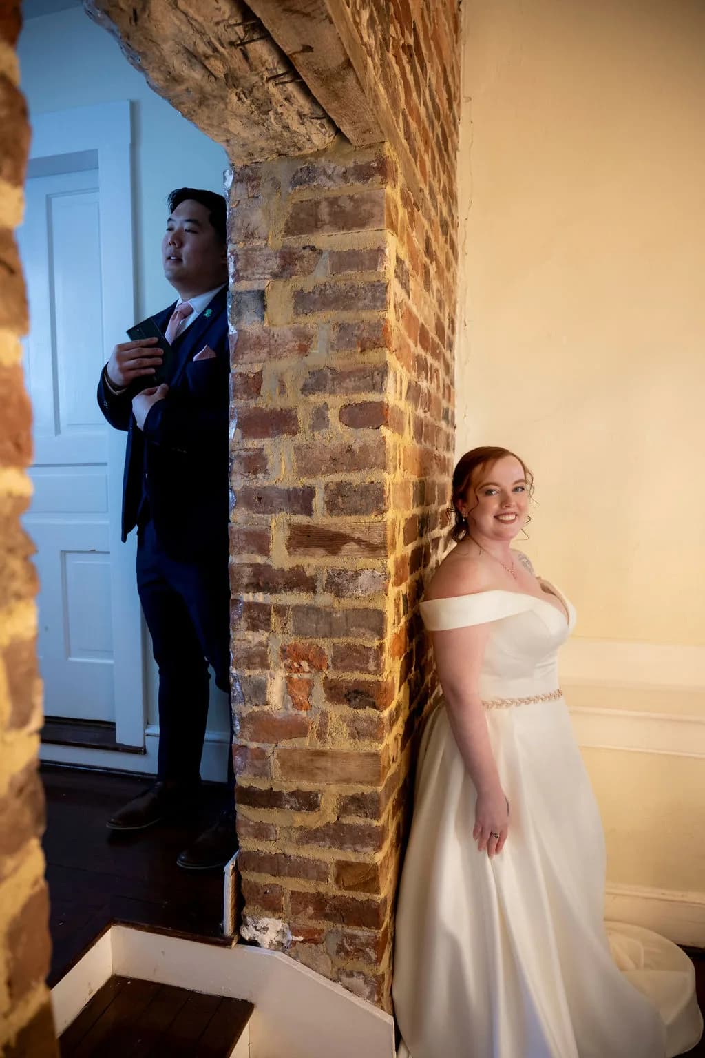 Bride and groom separated by a rustic brick pillar inside Rixey Manor, bride smiling at camera