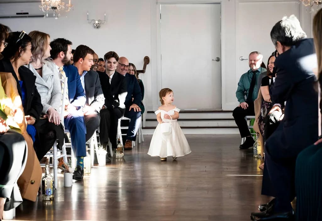 Tiny flower girl walks the aisle alone past smiling wedding guests in a bright white ceremony space