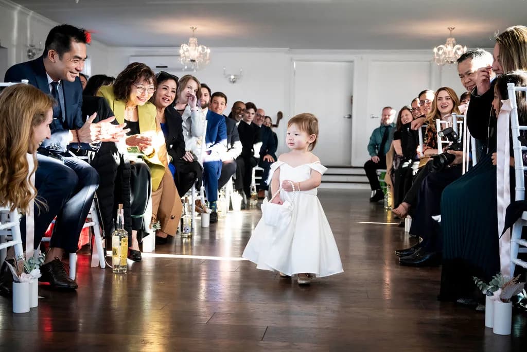 Toddler flower girl walks down wedding aisle in white dress as delighted guests smile and applaud