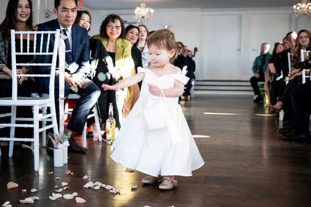 Tiny flower girl in white dress scatters petals down the aisle as delighted wedding guests look on