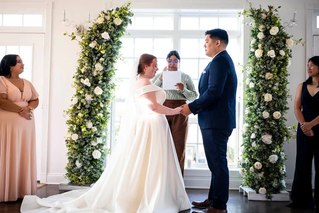 Bride and groom exchange vows beneath a lush floral arch with bridesmaids and officiant present