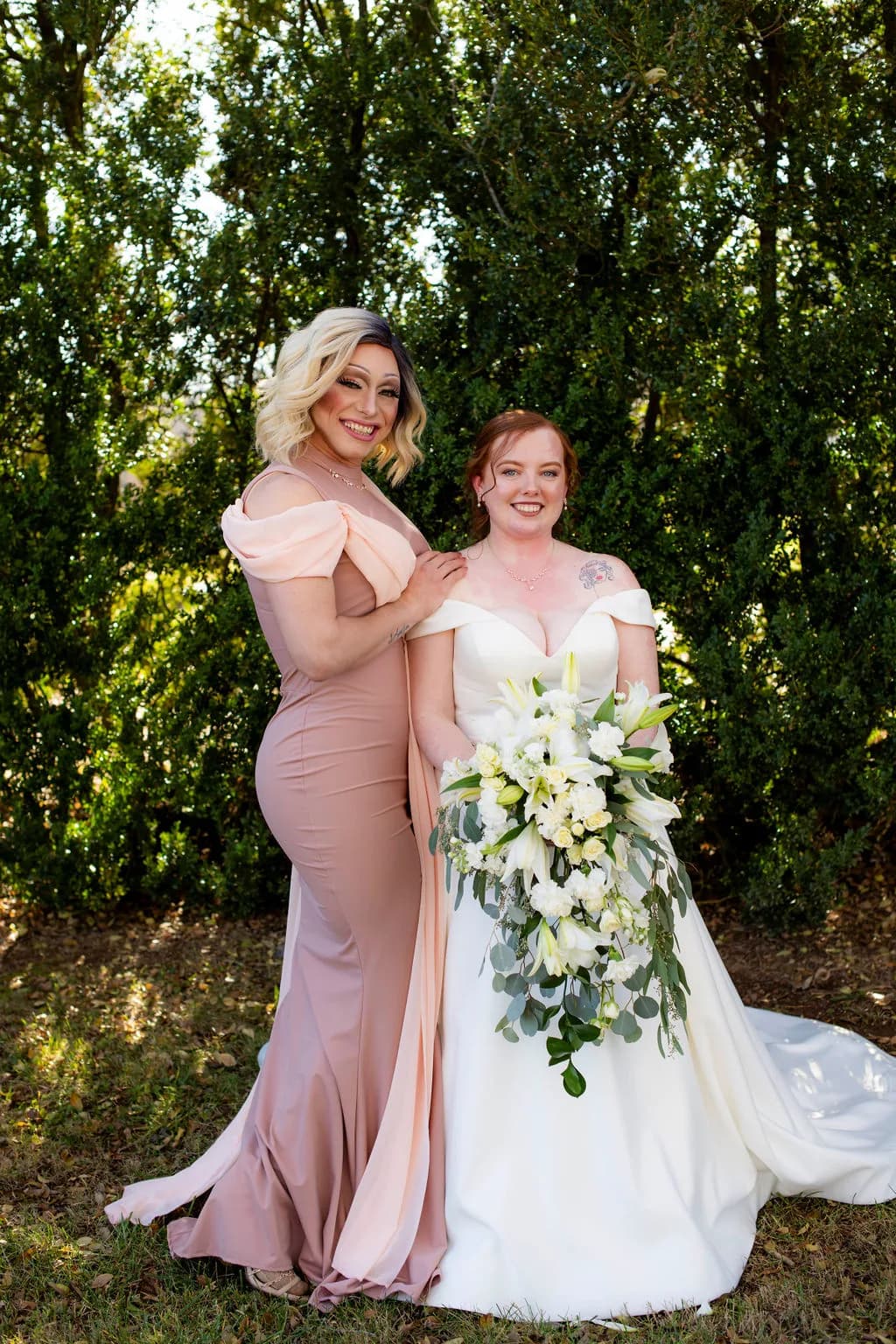 Smiling bride in white gown holds cascading white floral bouquet beside bridesmaid in blush dress outdoors