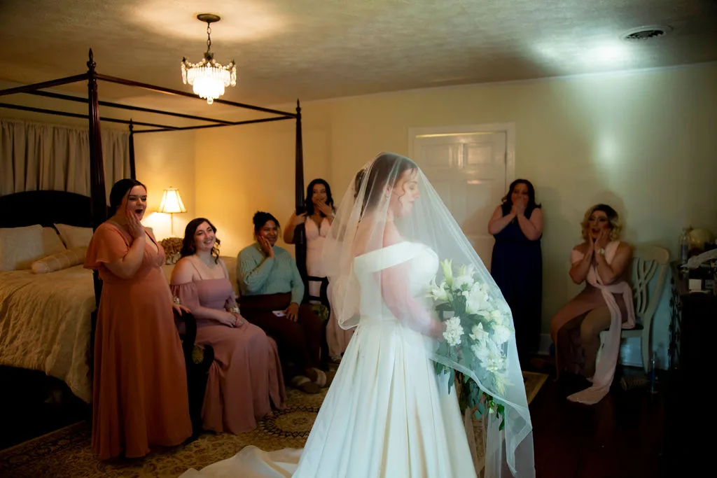 Bridesmaids react with awe and tears as bride reveals her wedding dress in a warmly lit bridal suite
