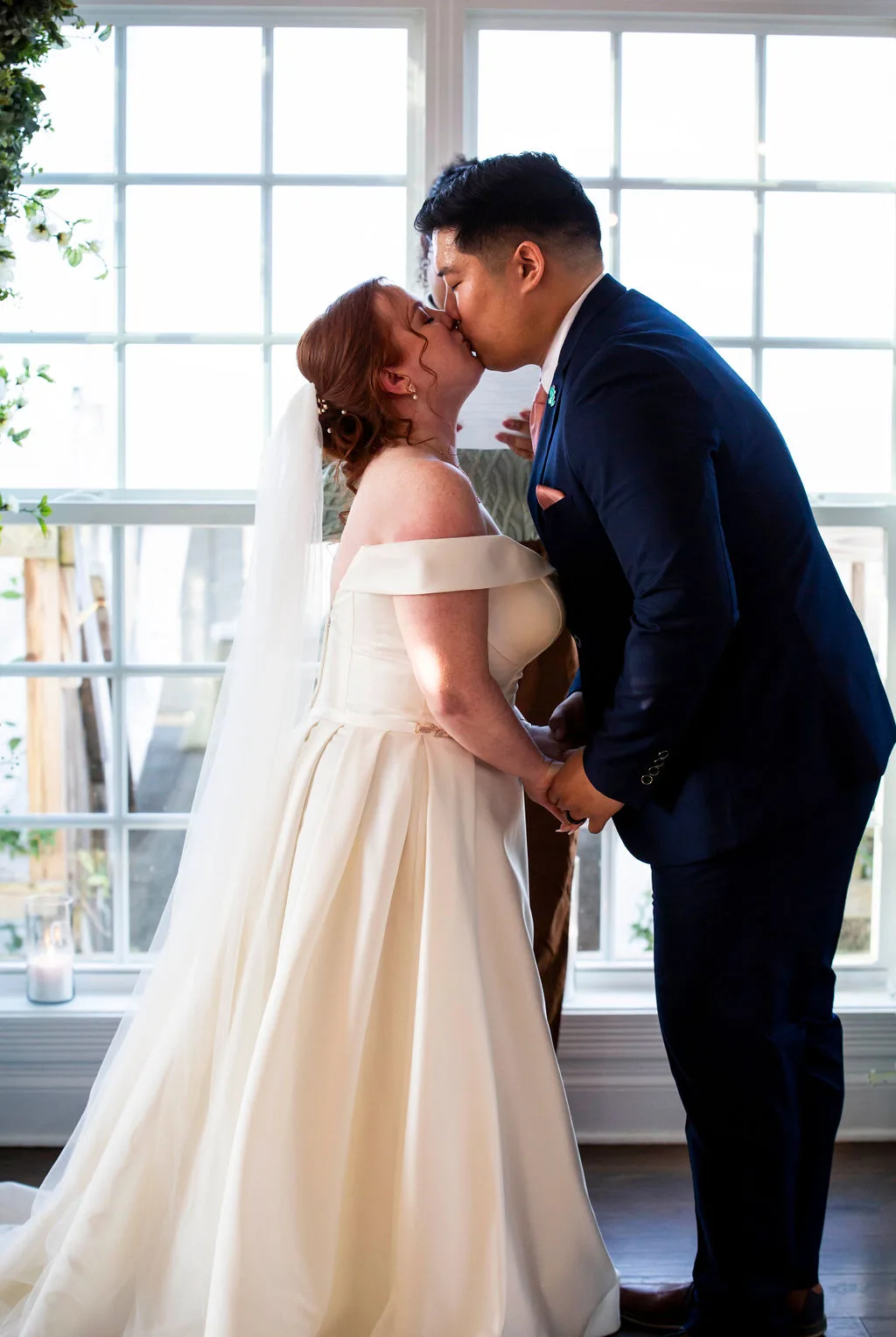 Bride and groom share first kiss at the altar, backlit by large windows at Rixey Manor ceremony