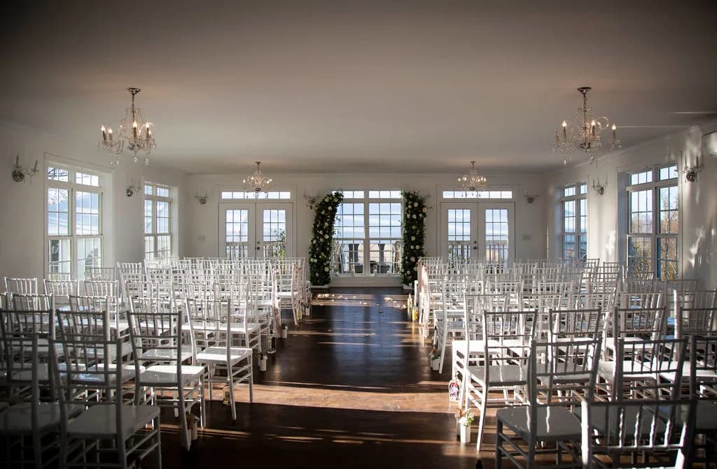 Sunlit ceremony hall at Rixey Manor with white chiavari chairs, floral arch, chandeliers, and wood floors
