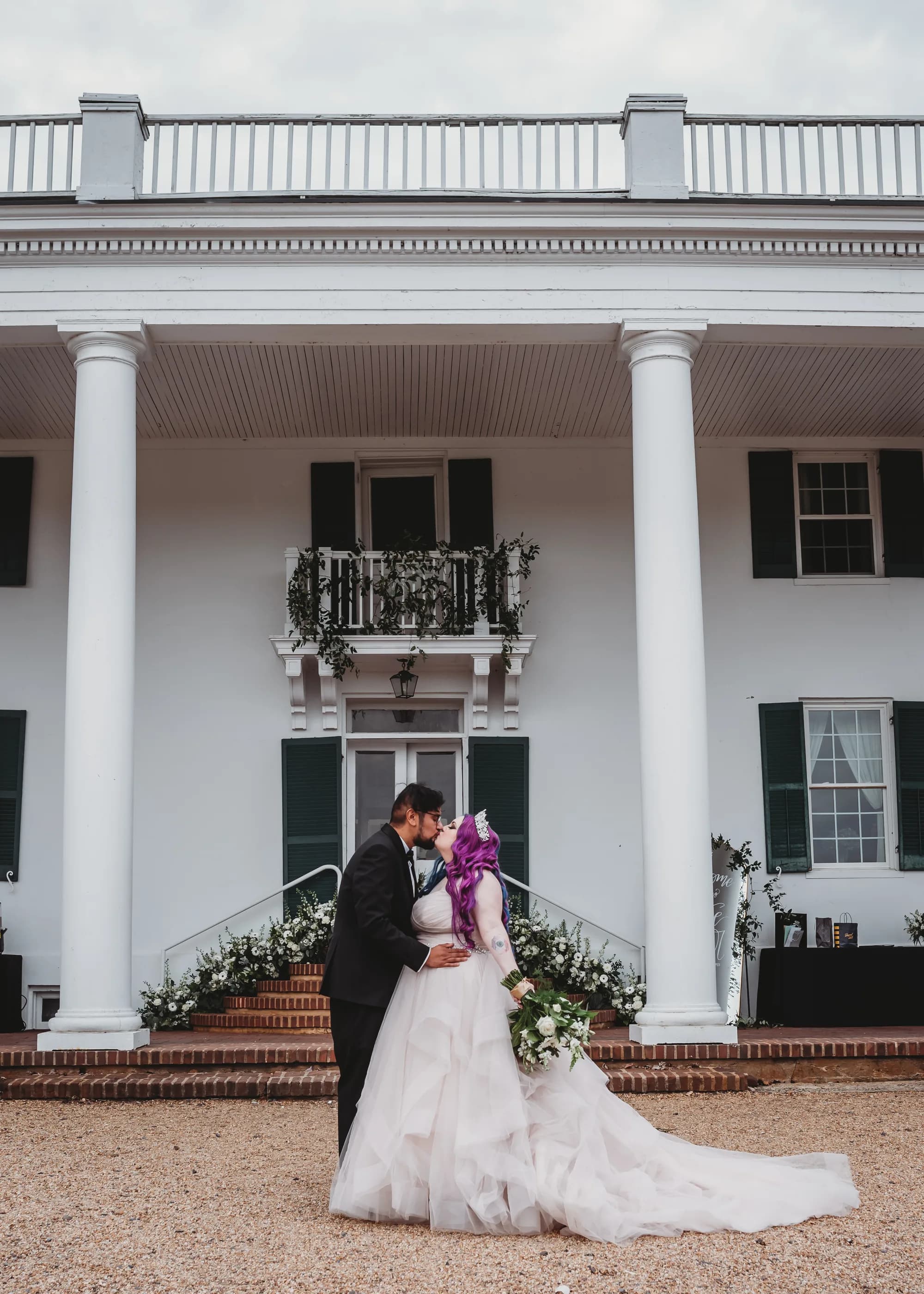 Bride with purple hair and groom kiss on gravel drive before Rixey Manor's white columned facade