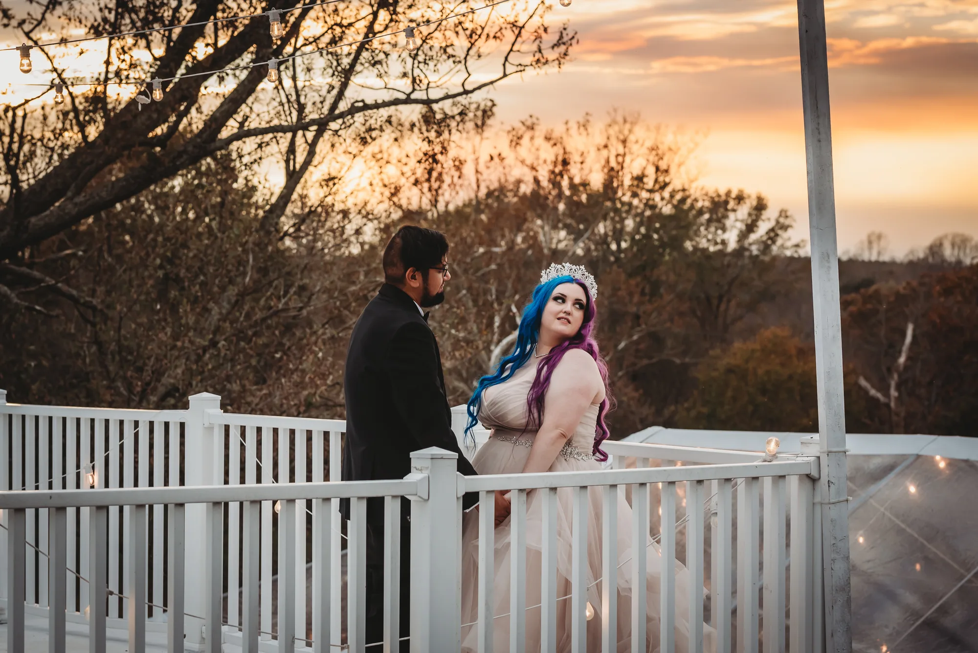 Bride with blue hair and tiara holds groom's hand on Rixey Manor deck at golden hour sunset