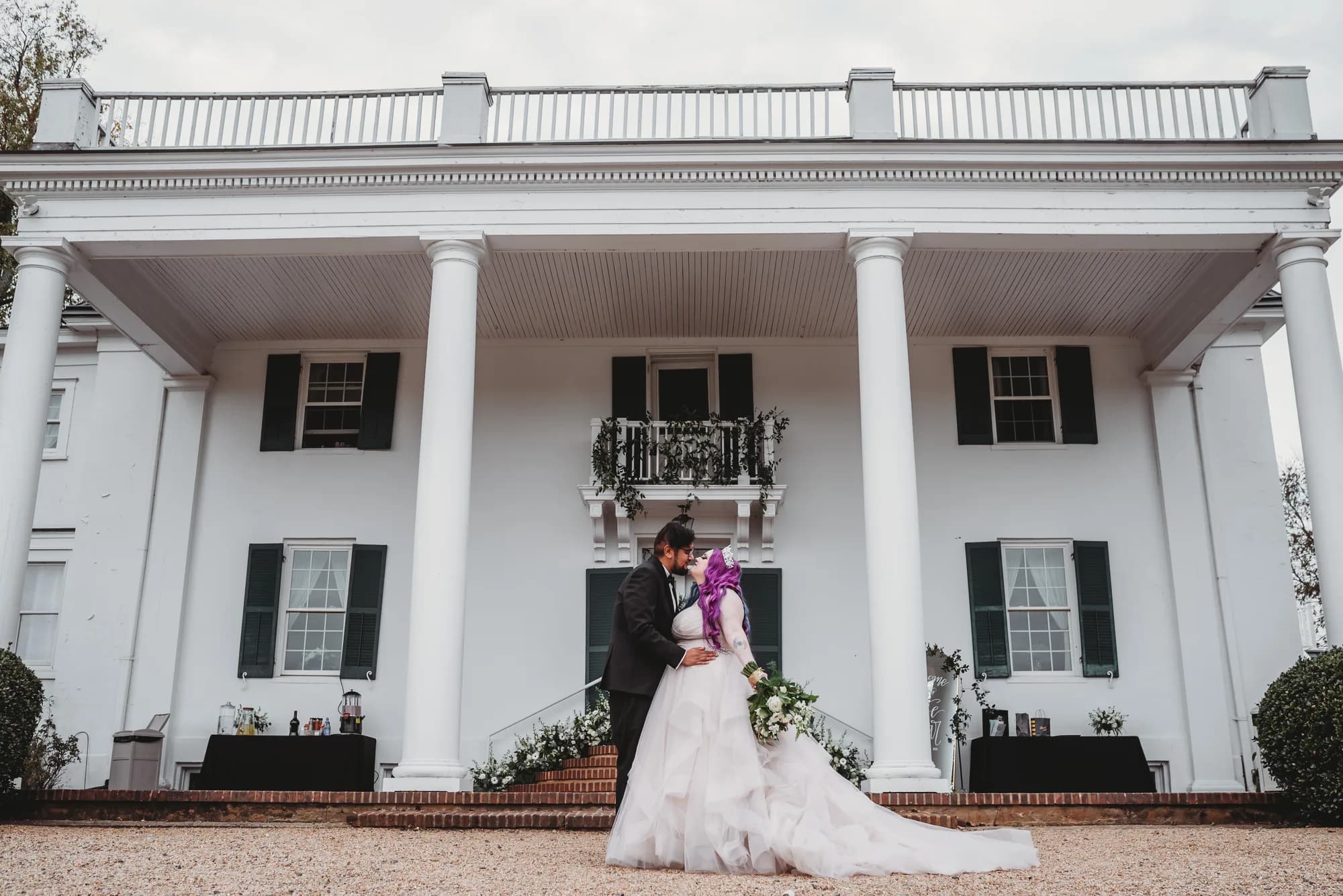 Bride and groom portrait in front of white columned manor house with brick walkway