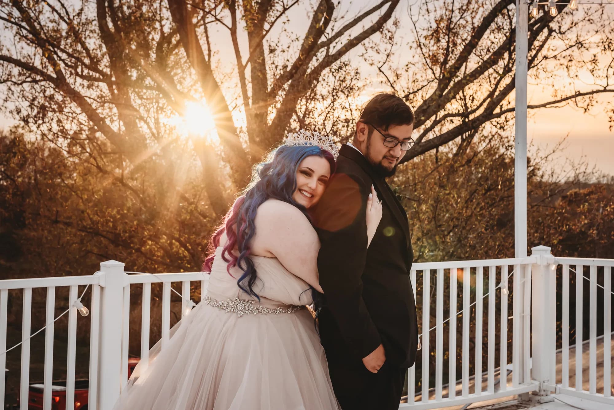 Bride with blue hair leans on groom at golden hour on Rixey Manor terrace, sun flaring through autumn trees