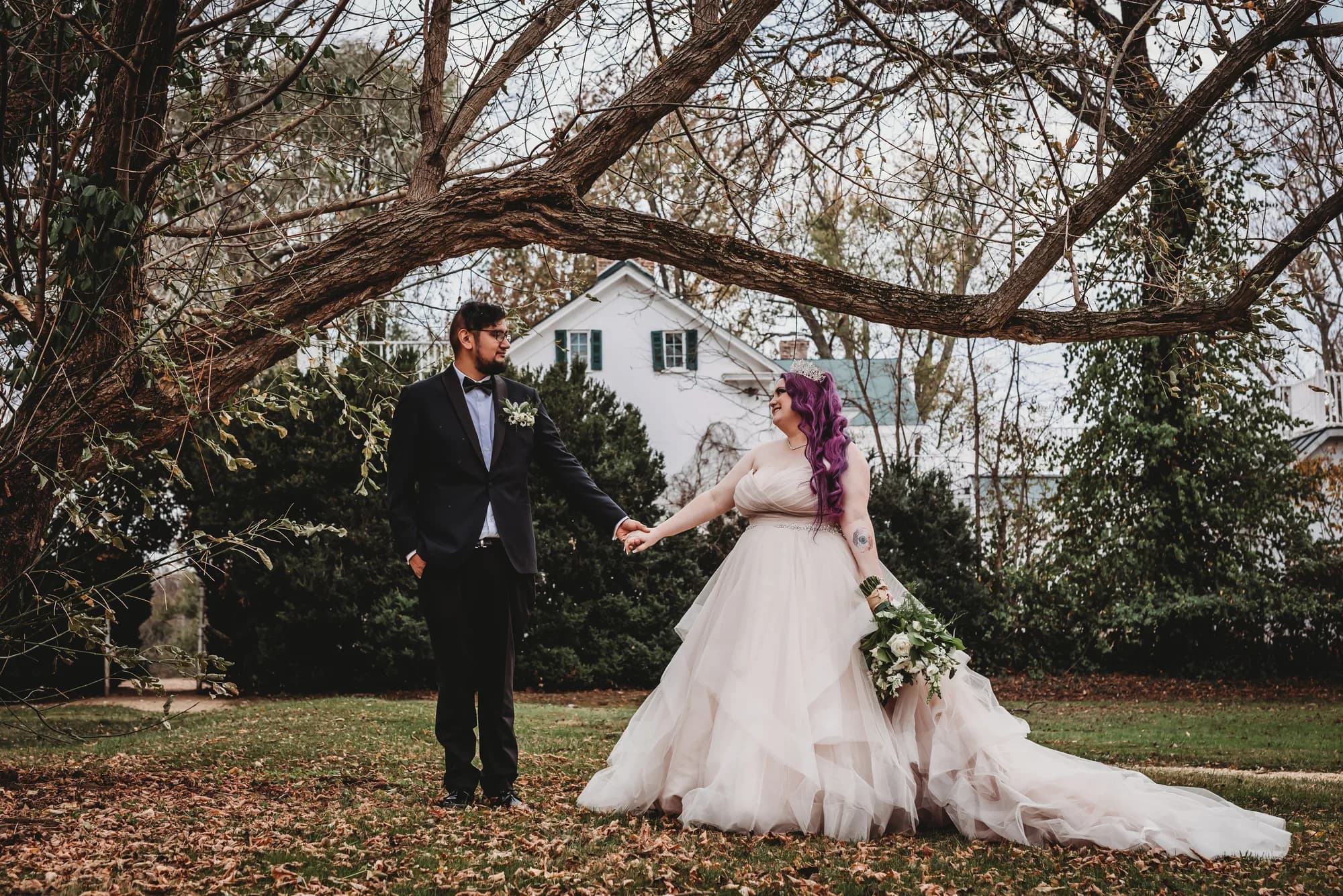Bride in layered gown and groom in tuxedo hold hands under bare autumn trees on Rixey Manor grounds