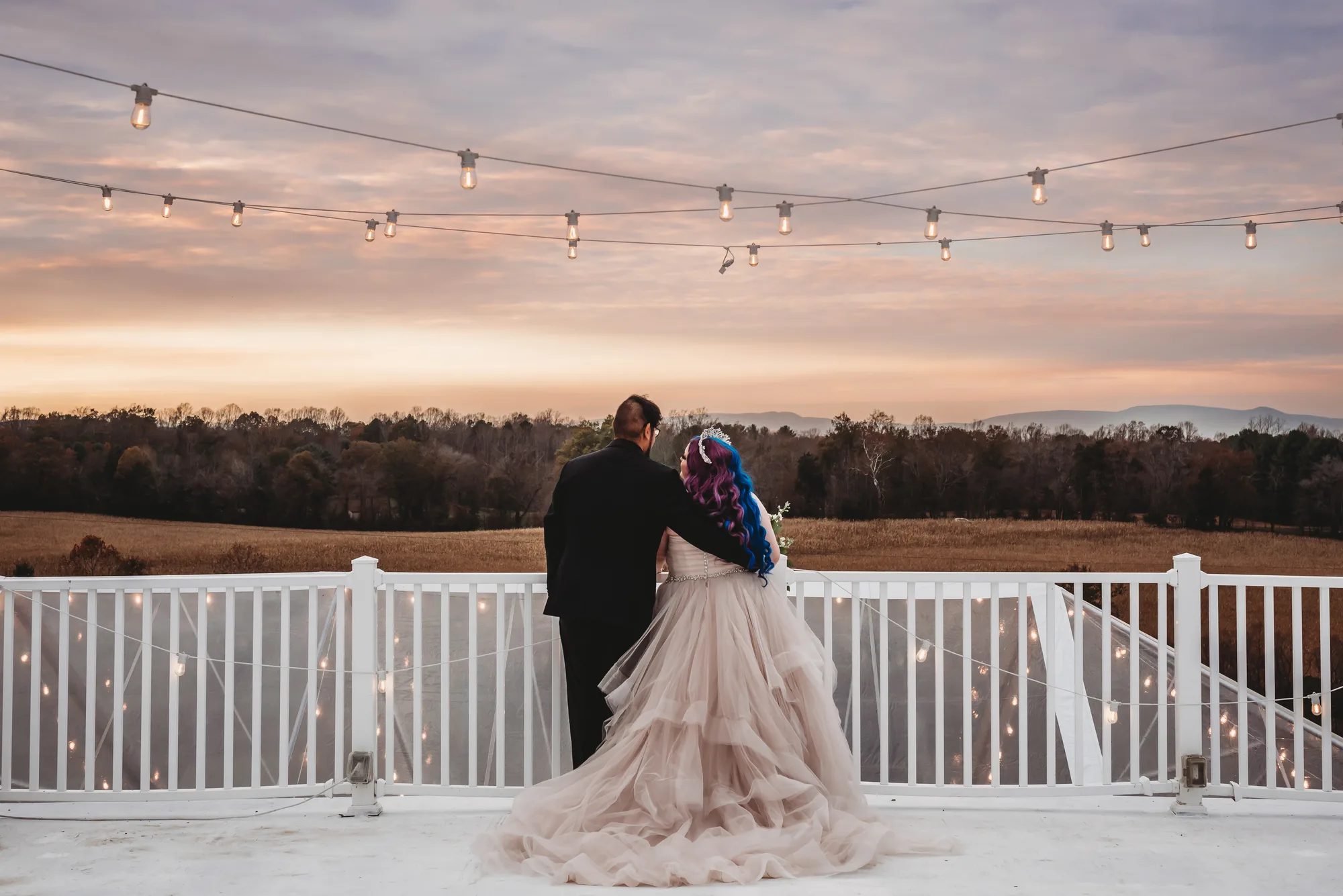 Bride and groom overlook golden fields at sunset from Rixey Manor rooftop deck under string lights