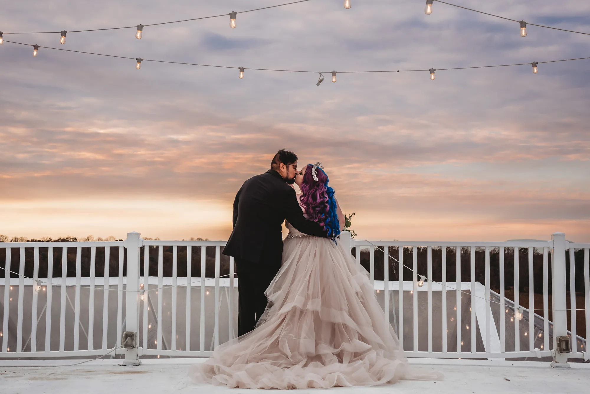 Couple sharing first kiss on Rixey Manor rooftop terrace under string lights at dramatic sunset