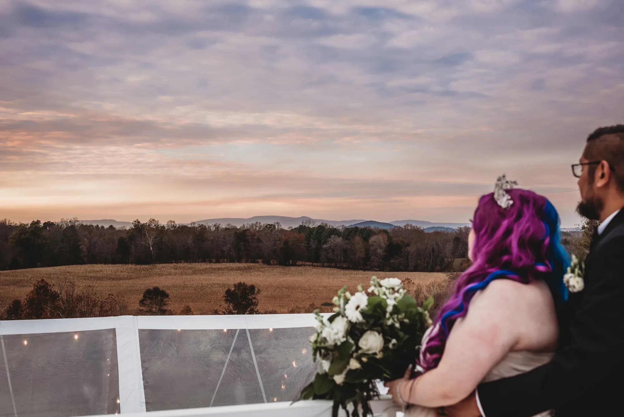 Couple with purple-haired bride holding bouquet overlooks Virginia countryside at sunset from Rixey Manor terrace