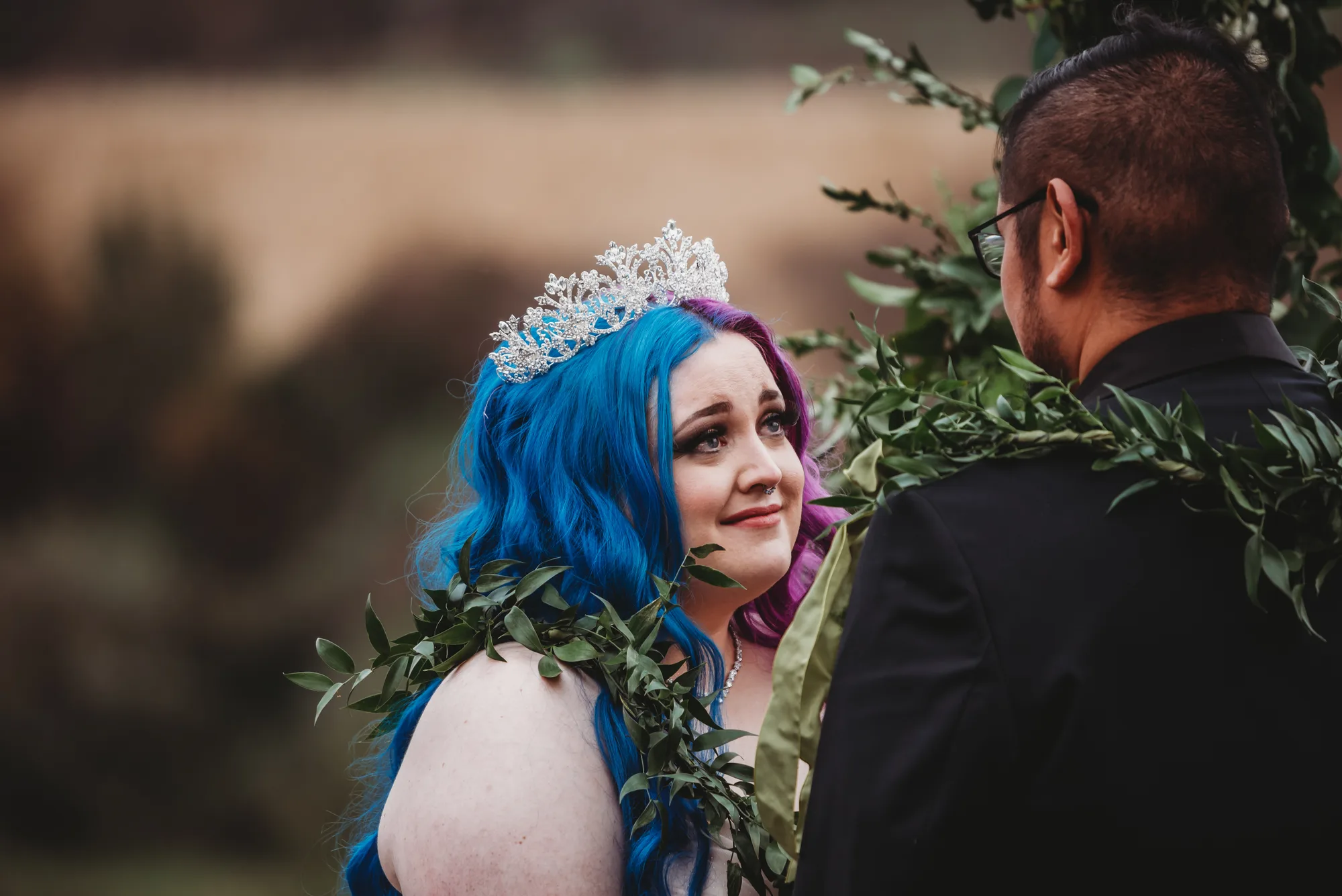 Bride with blue hair and tiara gazes at groom during outdoor ceremony beneath greenery arch