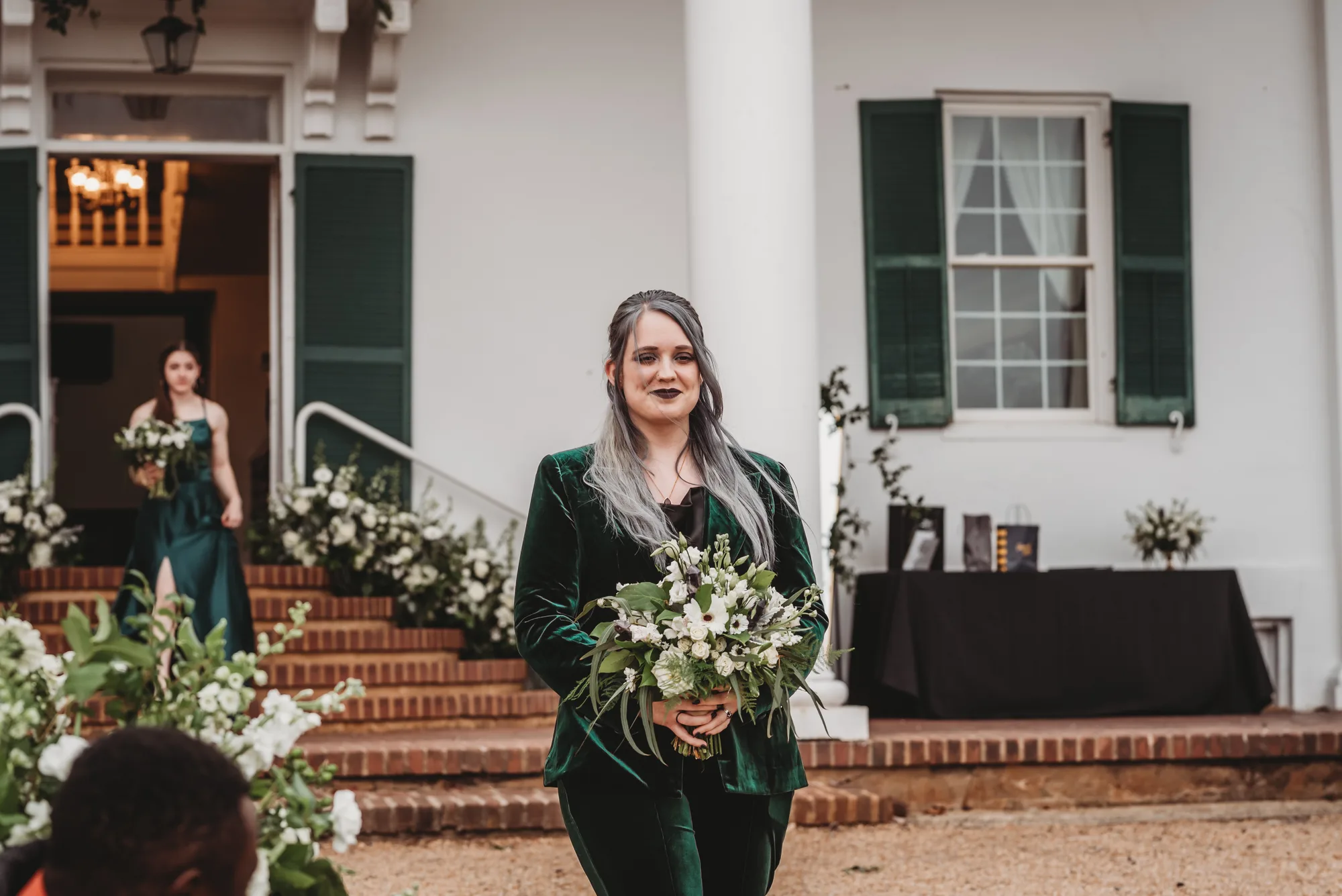 Bridesmaid in emerald velvet suit walks aisle holding white bouquet at Rixey Manor's columned estate entrance
