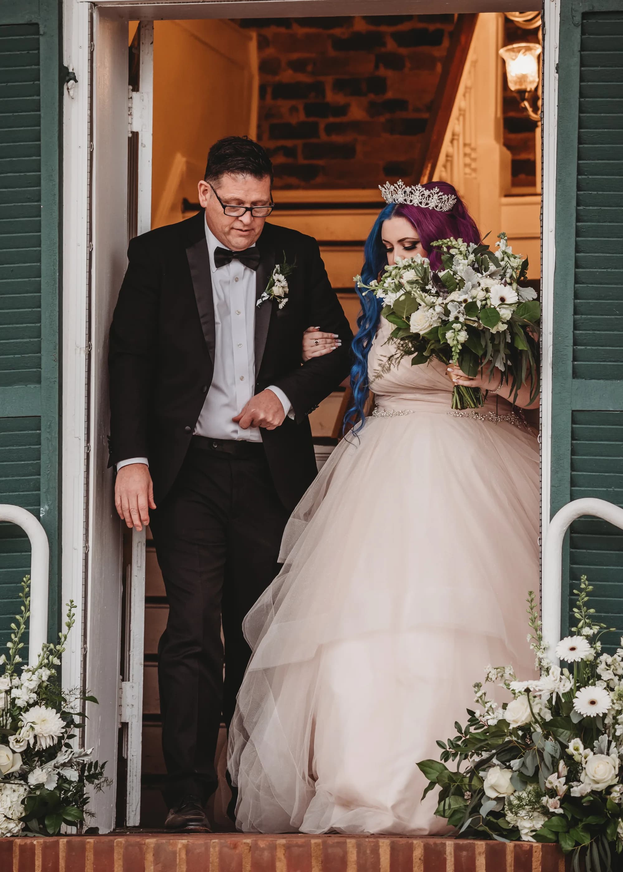 Bride with colorful hair and tiara exits Rixey Manor doors with escort, carrying white floral bouquet