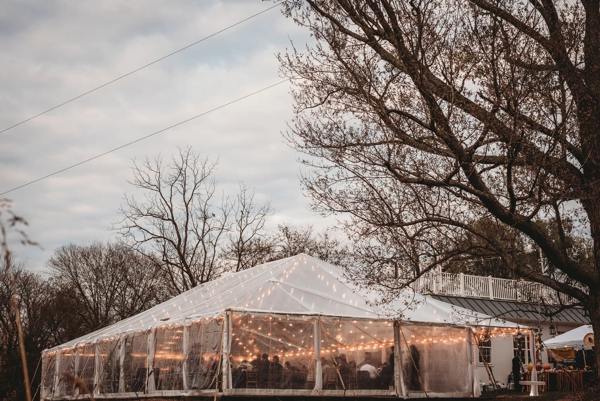 Glowing transparent reception tent draped in string lights at dusk among bare trees at Rixey Manor