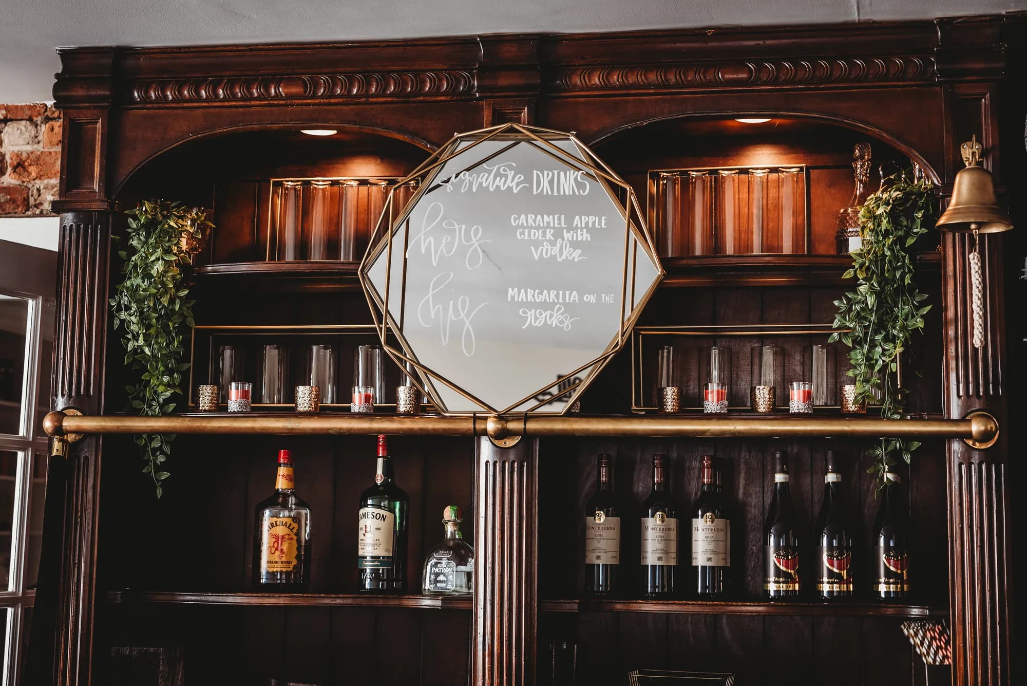 Dark wood bar at Rixey Manor with signature cocktail mirror sign, liquor bottles, and lush greenery accents