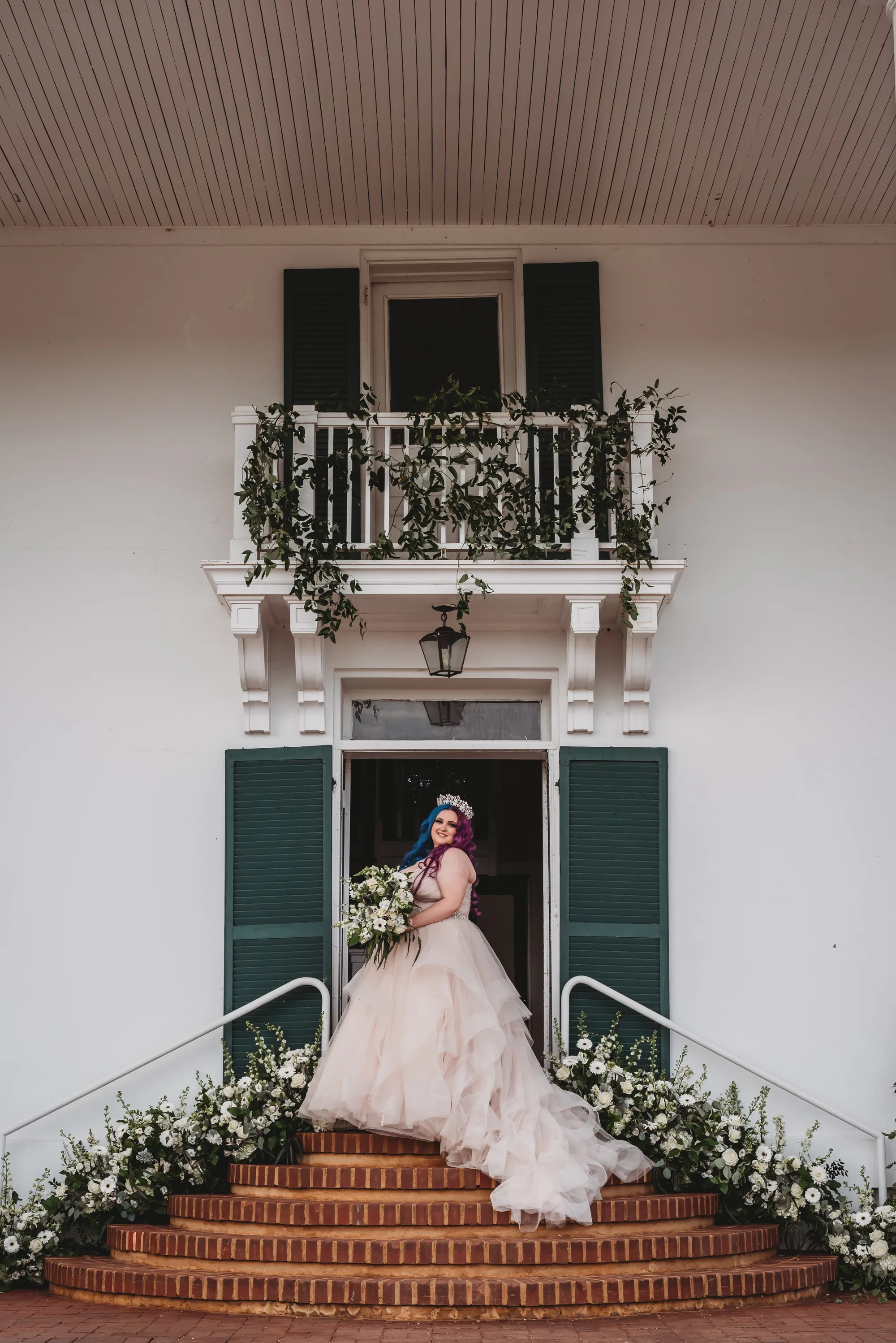Bride with purple hair in layered gown poses at Rixey Manor's white colonial entrance with floral-lined brick steps