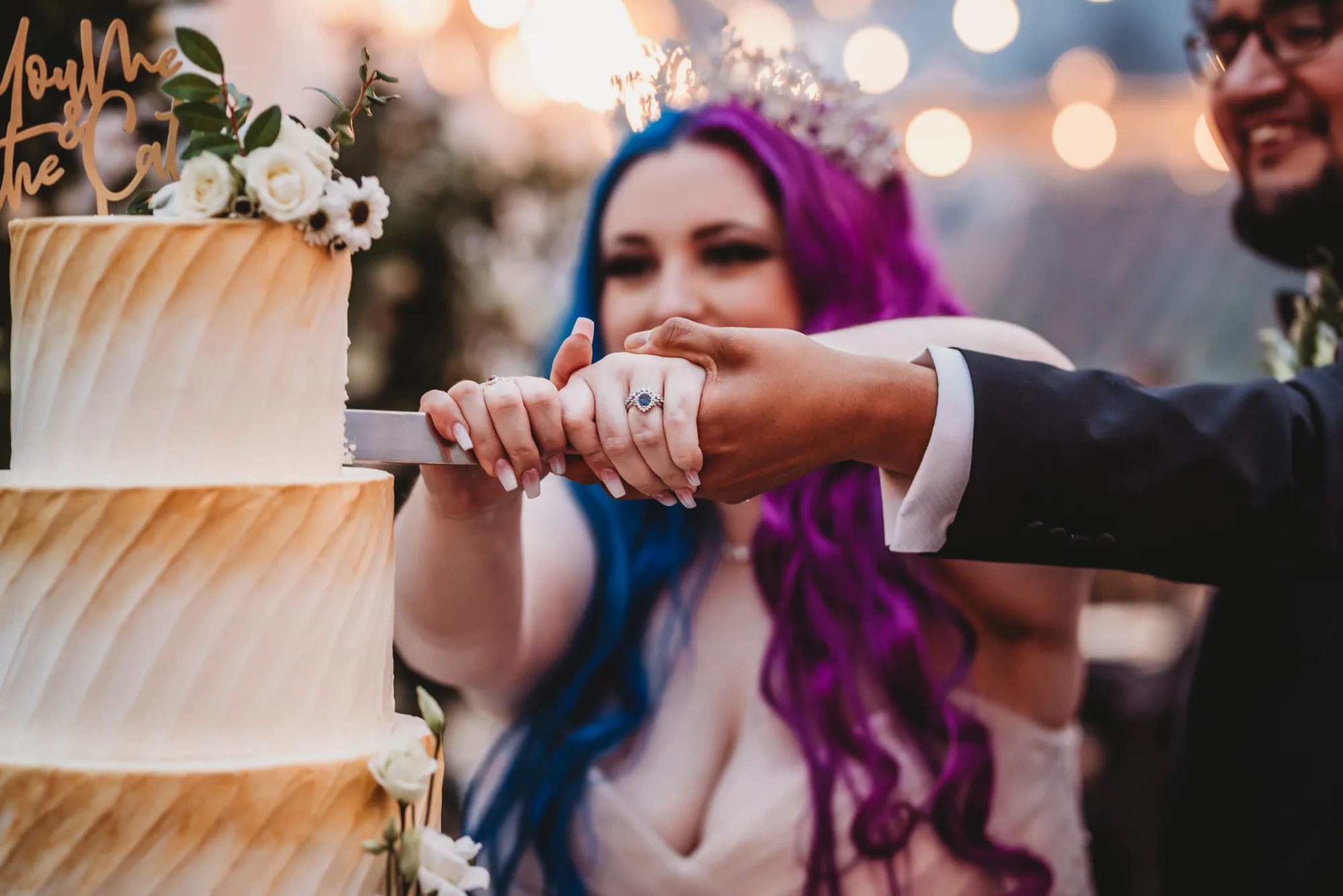 Bride with vivid purple and blue hair cuts wedding cake with groom, ring detail prominent, warm bokeh lights behind