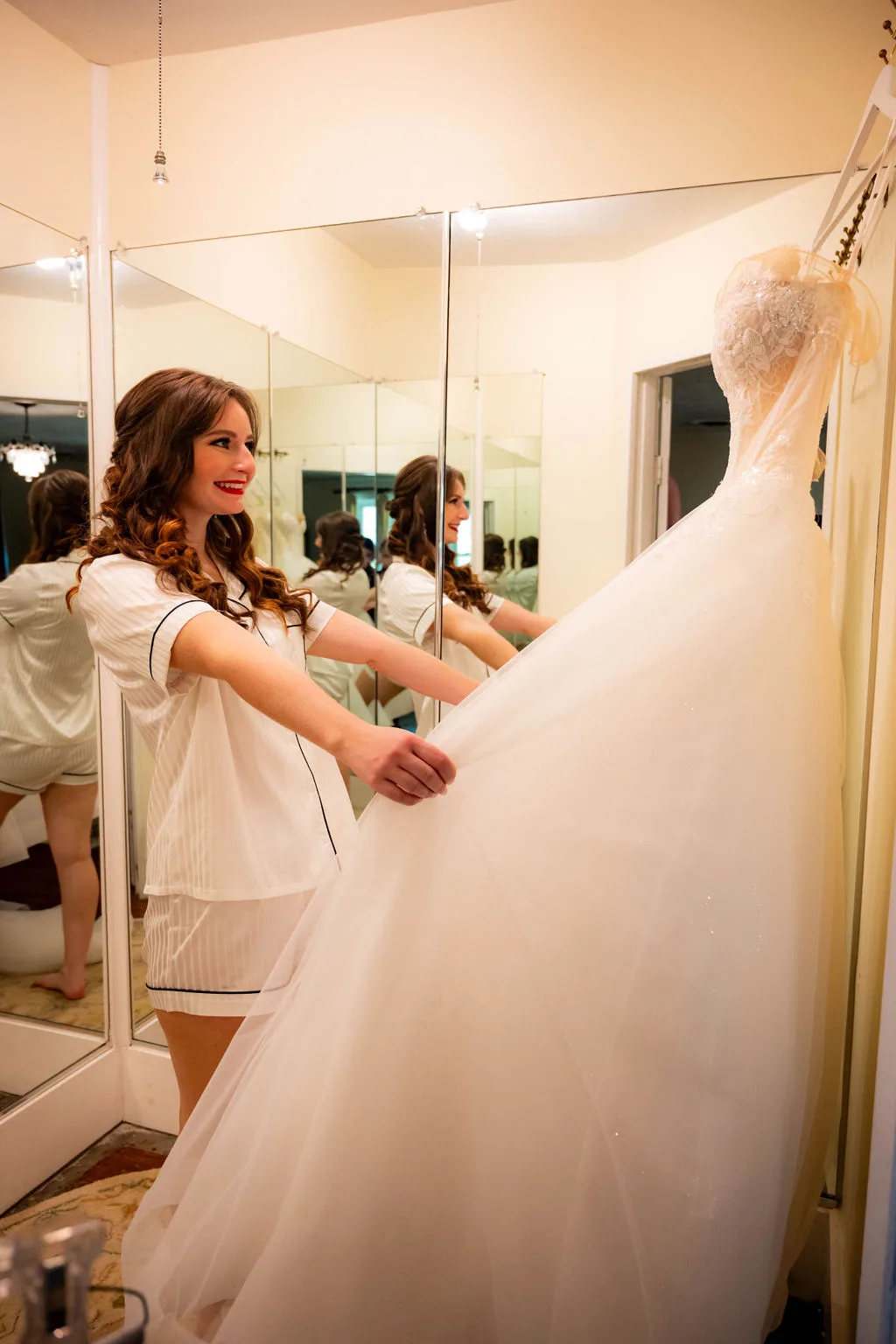 Smiling bride in pajamas holding her wedding gown in a mirrored dressing room before the ceremony