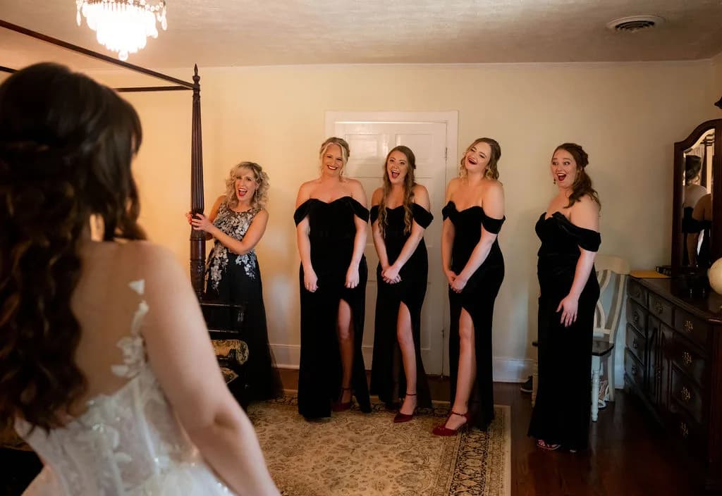 Bridesmaids in black gowns react with joy seeing bride in wedding dress during getting-ready moment