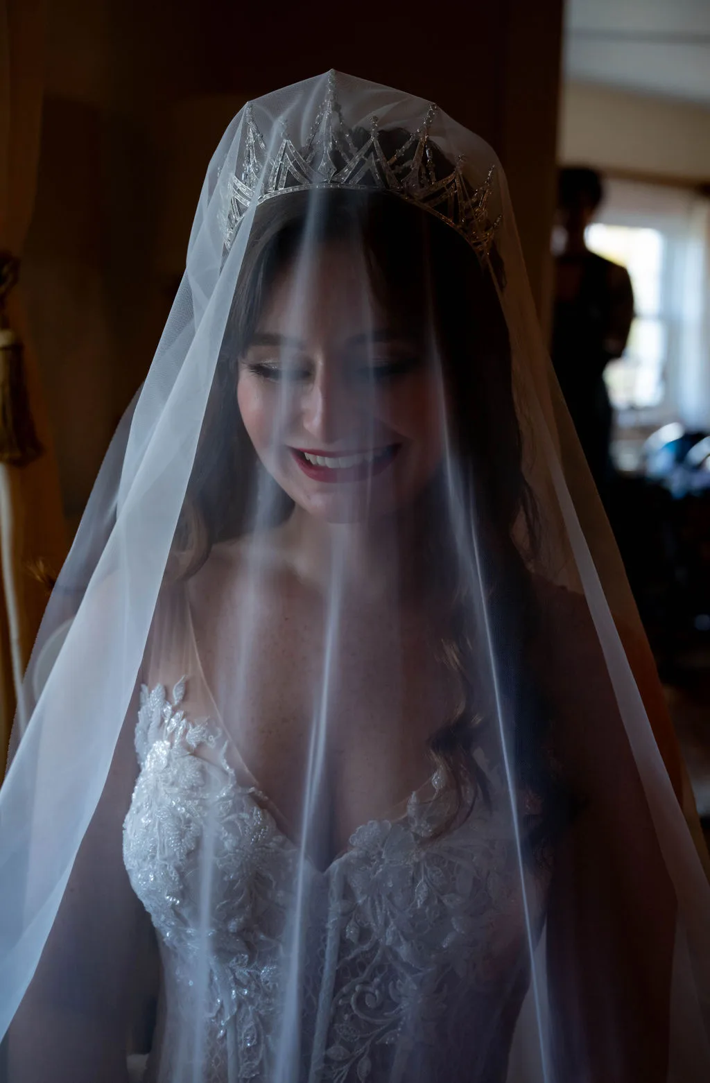 Bride smiling softly beneath sheer veil and crystal tiara in a warmly lit getting-ready room