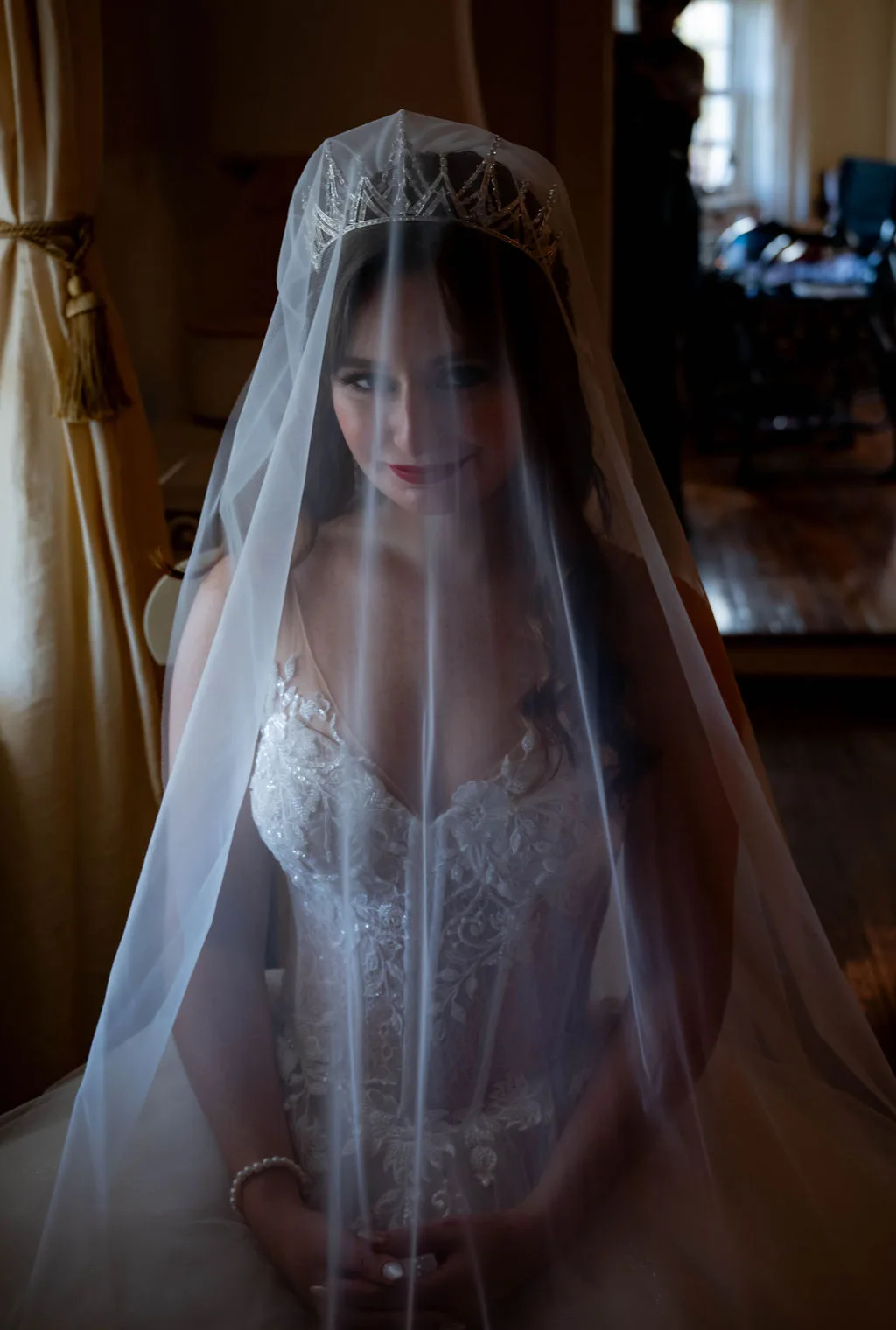 Bride in lace gown and crystal tiara smiles softly beneath her veil in a warmly lit manor room