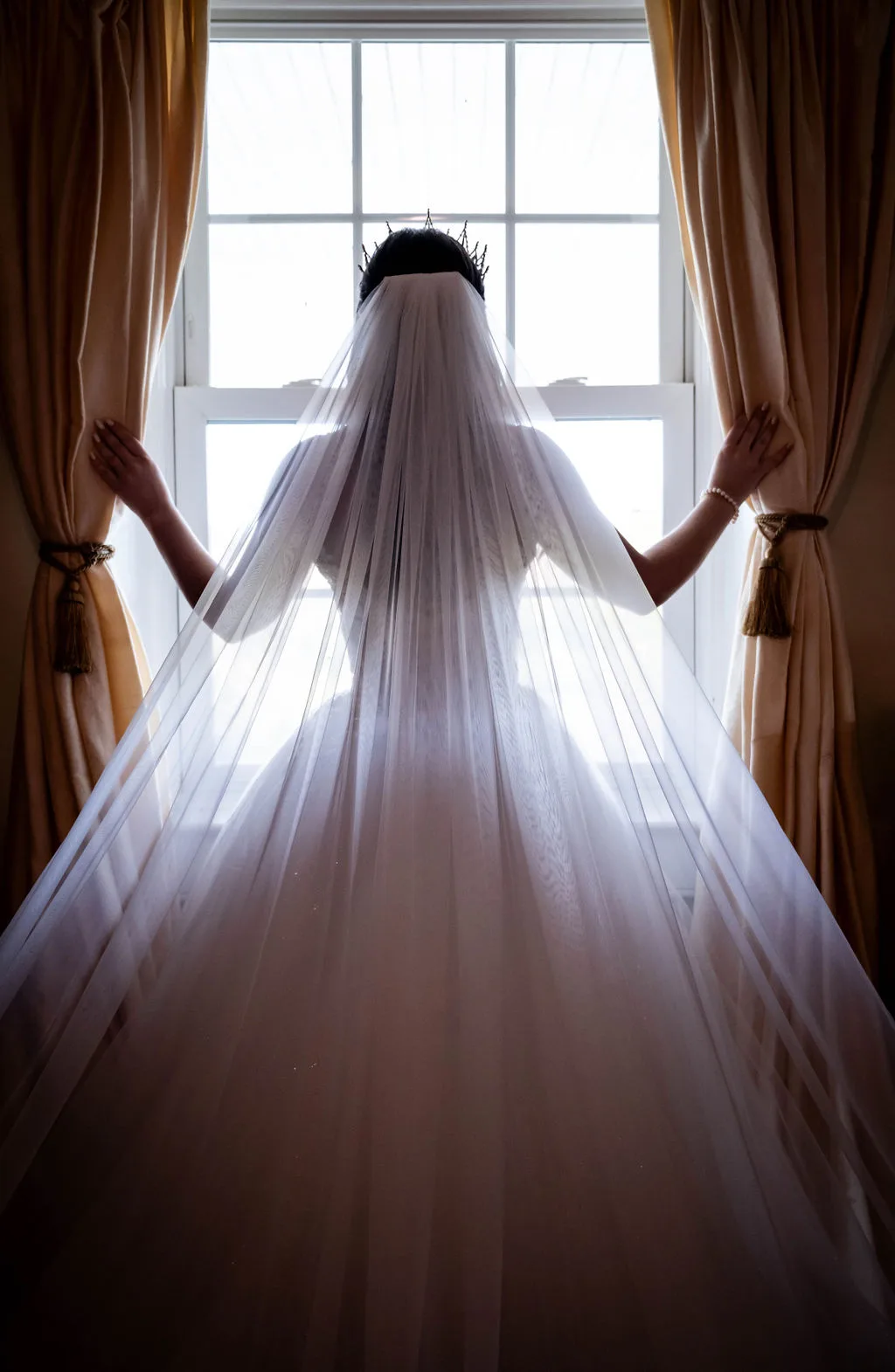 Bride in white veil and dress standing at window with arms outstretched, backlit by natural light
