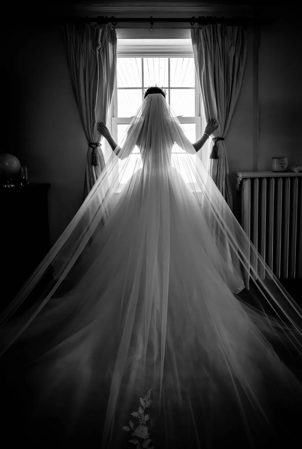 Bride in silhouette wearing cathedral veil and wedding gown, arms outstretched at sunlit window